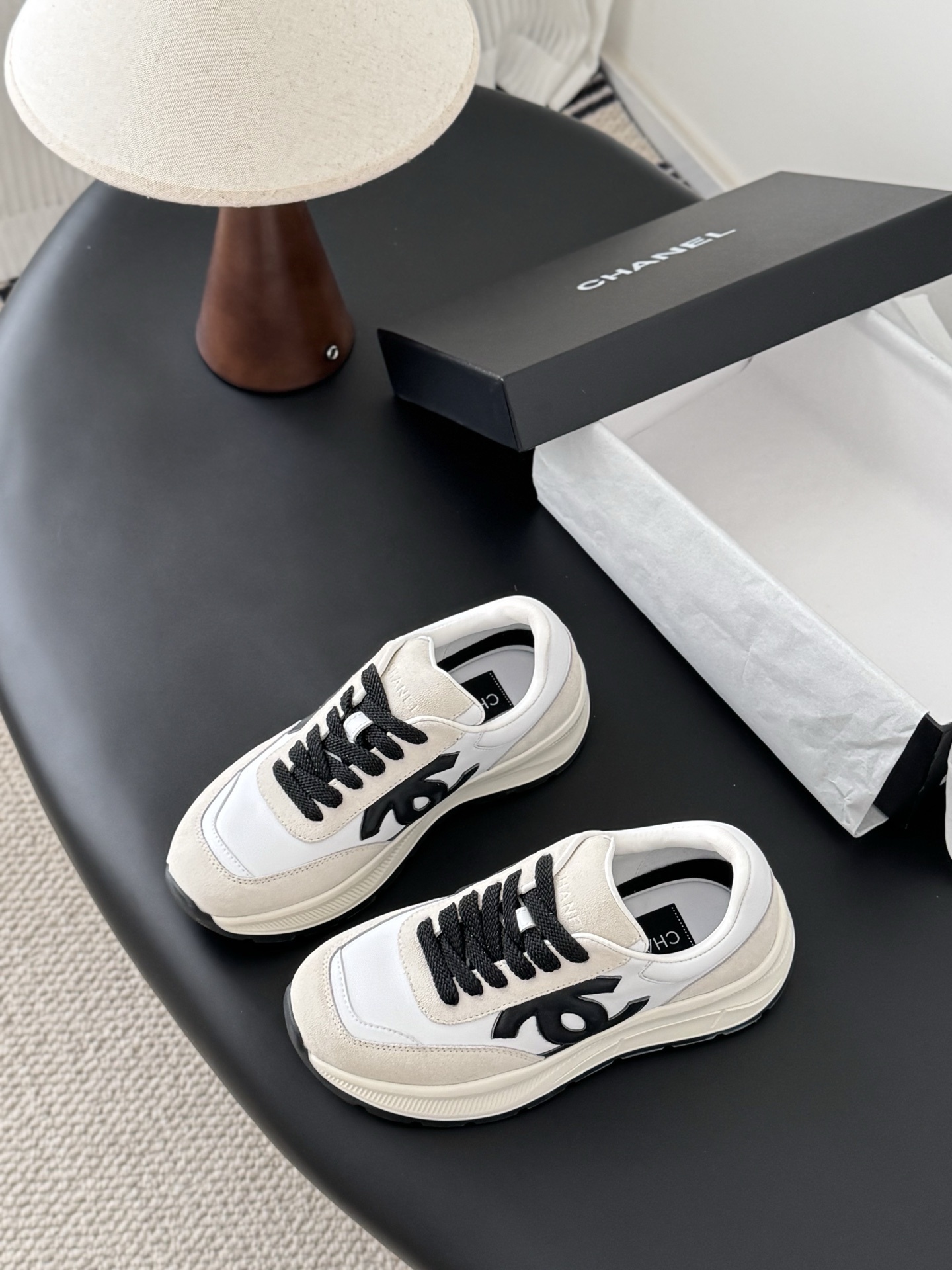 A pair of Chanel white and beige suede sneakers with black CC logo and laces displayed next to a black Chanel shoe box. - Hình 5