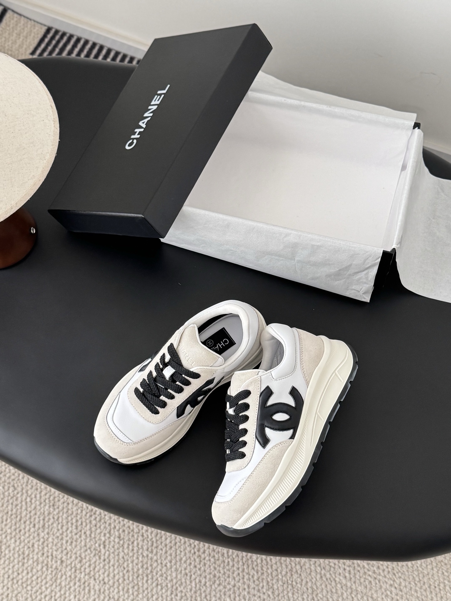 A pair of Chanel white and beige suede sneakers with black CC logo and laces displayed next to a black Chanel shoe box. - Hình 3