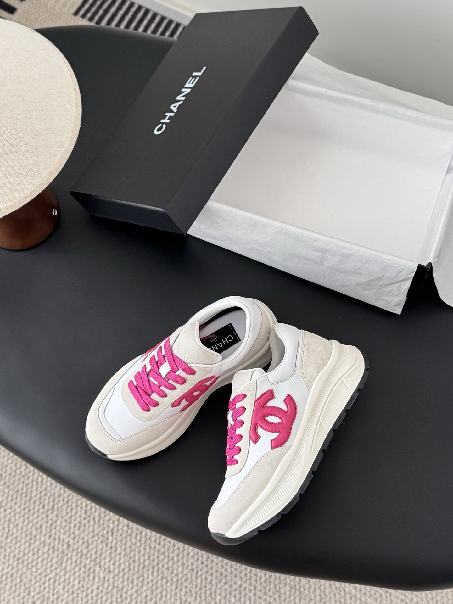 A pair of white and cream Chanel sneakers with bright pink laces and a pink CC logo placed next to a black Chanel shoebox. - Hình 3