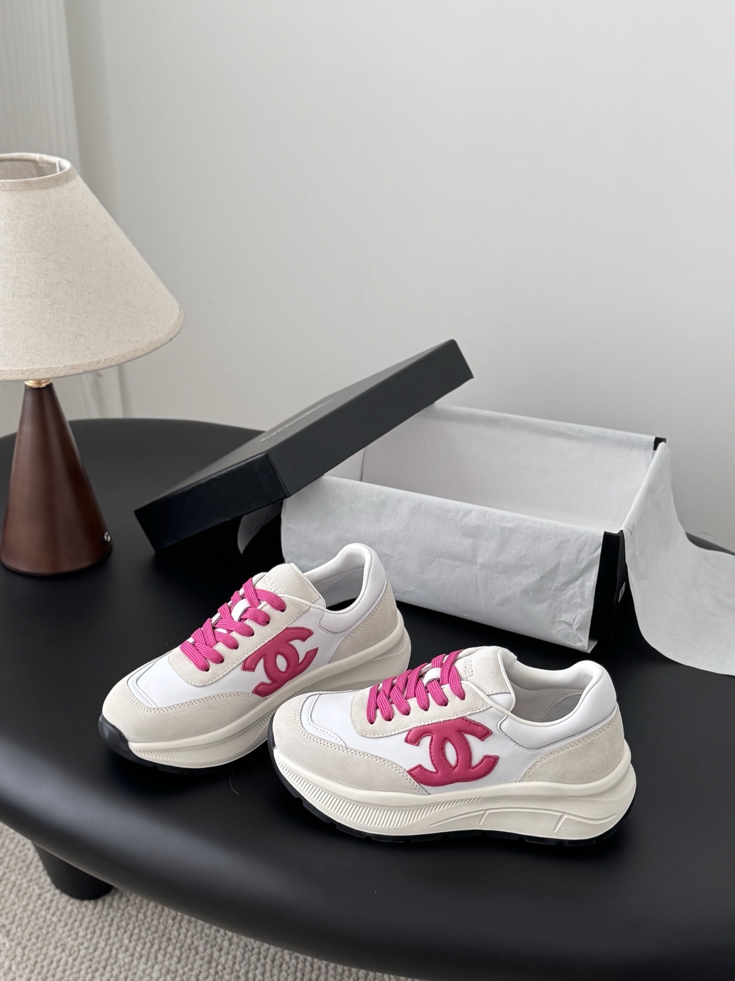 A pair of white and cream Chanel sneakers with bright pink laces and a pink CC logo placed next to a black Chanel shoebox. - Hình 2