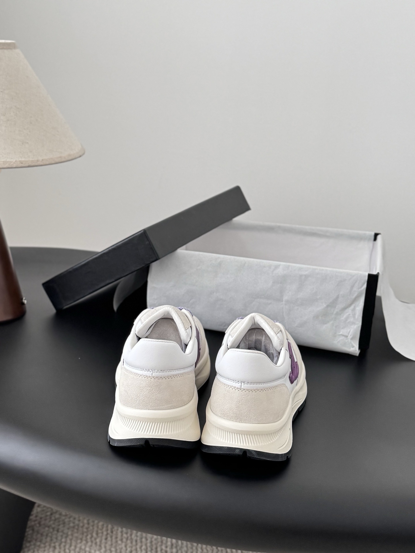 Pair of Chanel white leather sneakers with purple CC logo and purple laces on a black table next to a Chanel box. - Hình 7