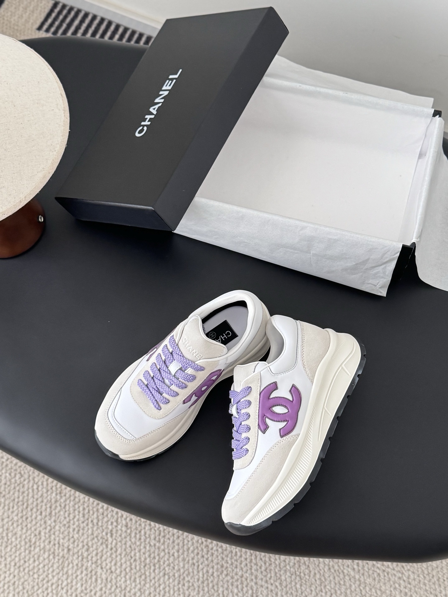 Pair of Chanel white leather sneakers with purple CC logo and purple laces on a black table next to a Chanel box. - Hình 3