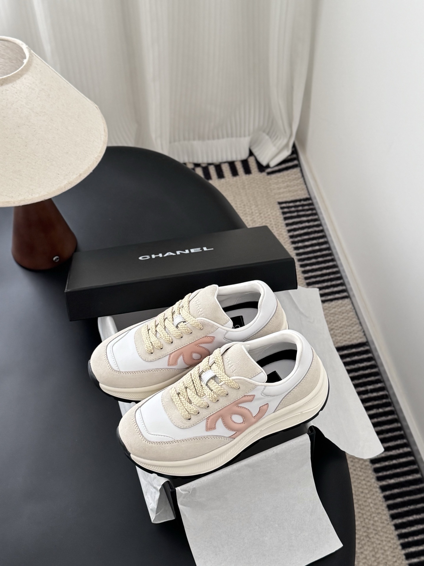 A pair of Chanel white and beige luxury sneakers with a soft pink CC logo sitting on a black surface next to an open Chanel shoe box. - Hình 9