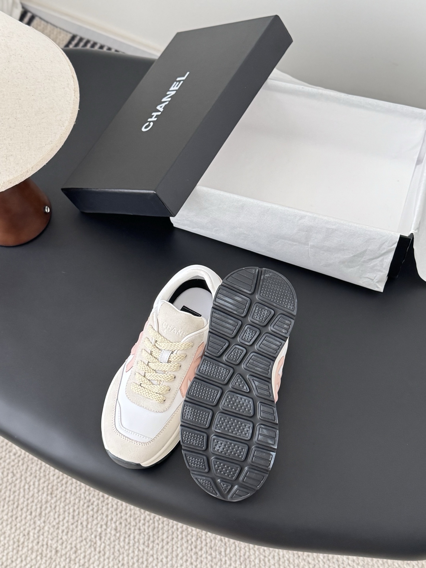 A pair of Chanel white and beige luxury sneakers with a soft pink CC logo sitting on a black surface next to an open Chanel shoe box. - Hình 4