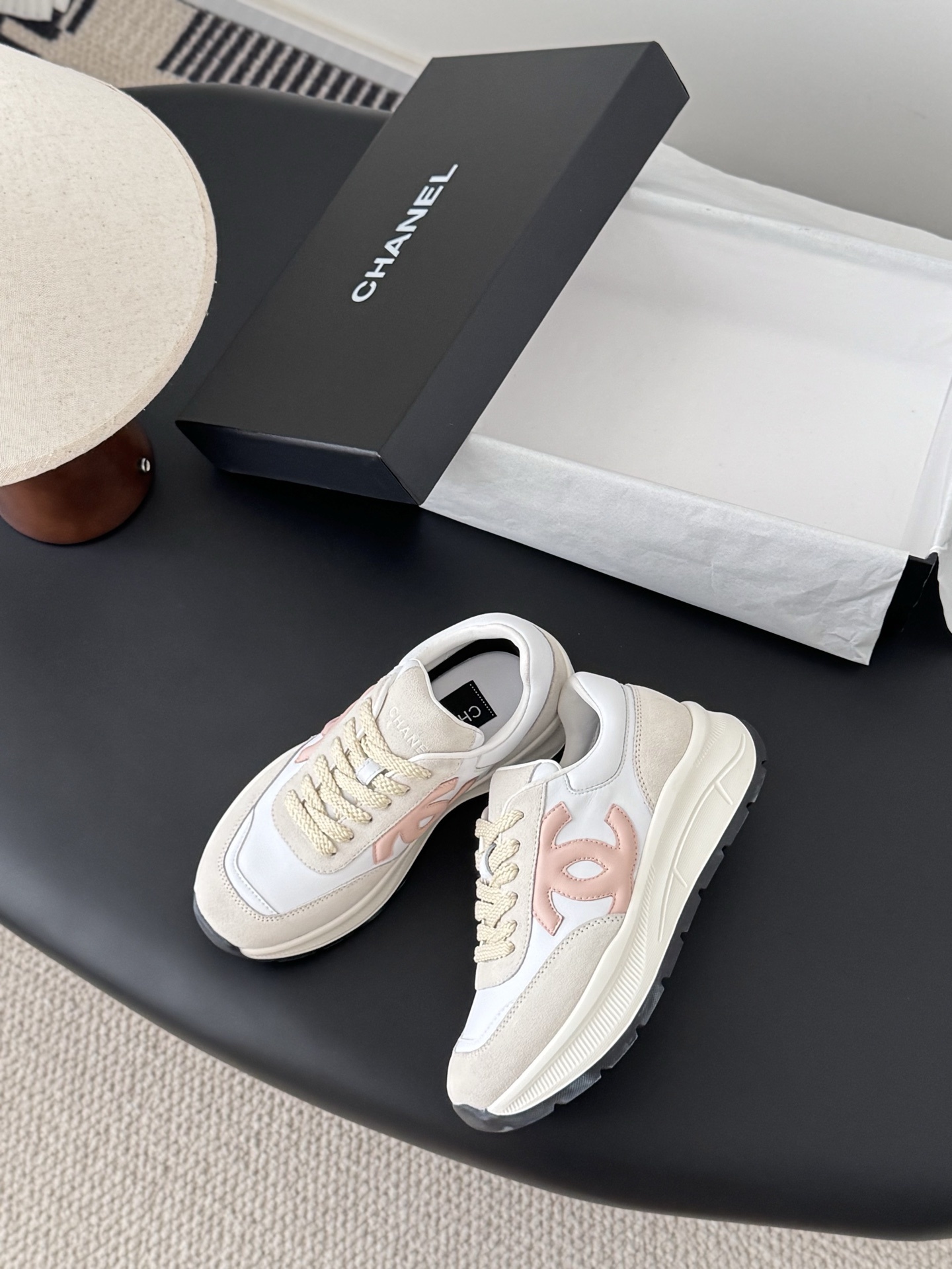 A pair of Chanel white and beige luxury sneakers with a soft pink CC logo sitting on a black surface next to an open Chanel shoe box. - Hình 3