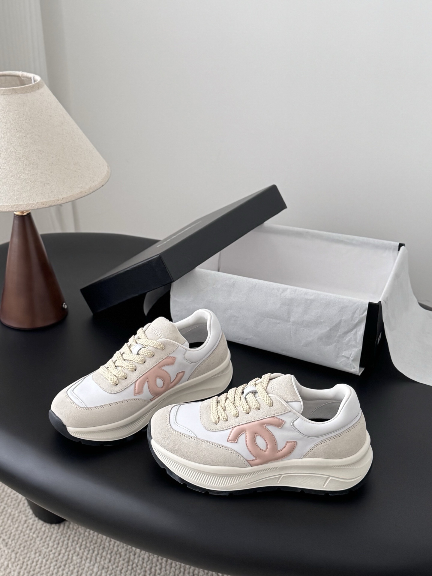 A pair of Chanel white and beige luxury sneakers with a soft pink CC logo sitting on a black surface next to an open Chanel shoe box. - Hình 2