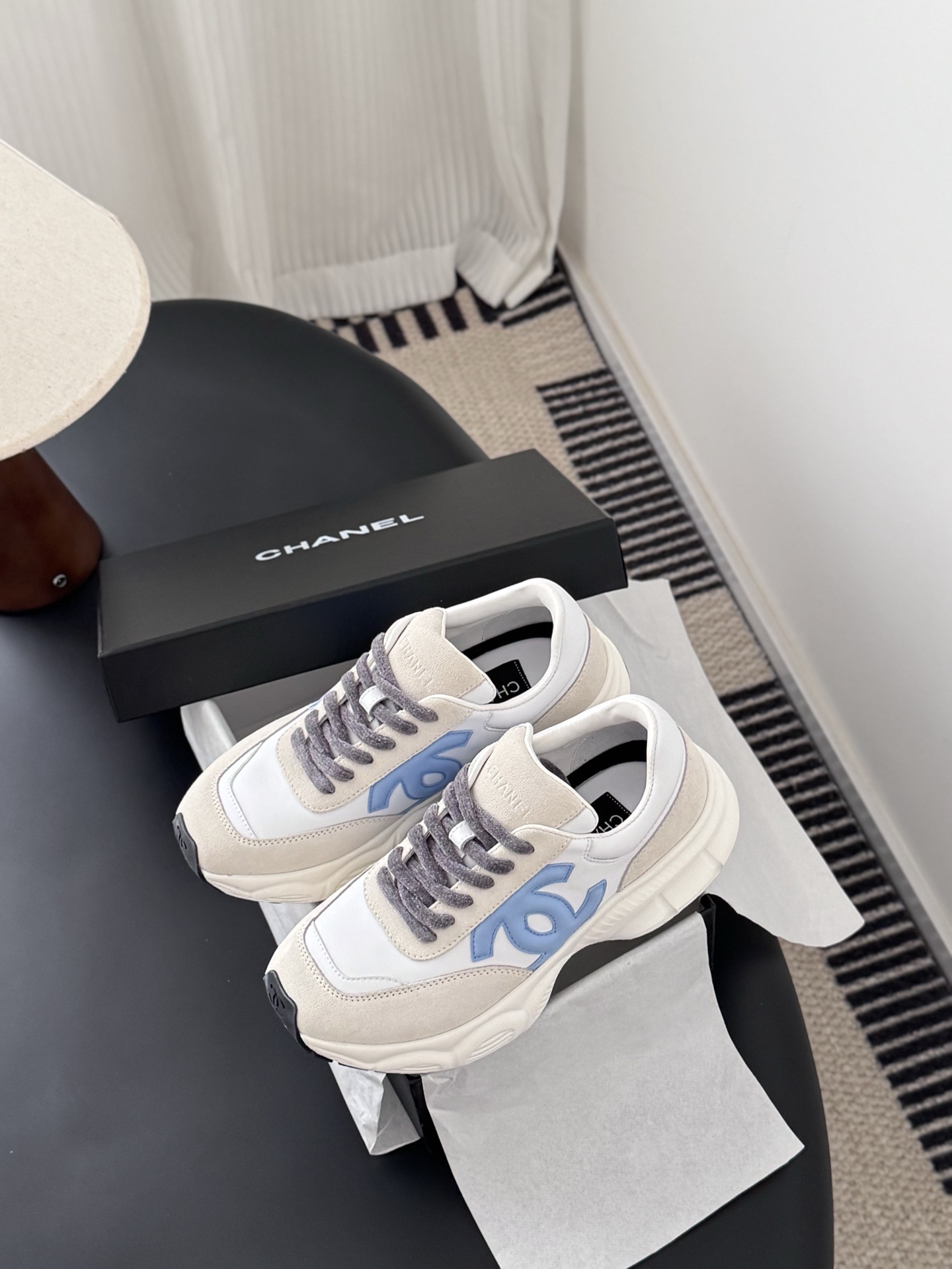 A pair of Chanel sneakers in white and beige suede with a light blue CC logo and grey laces, shown with a black Chanel shoebox. - Hình 9