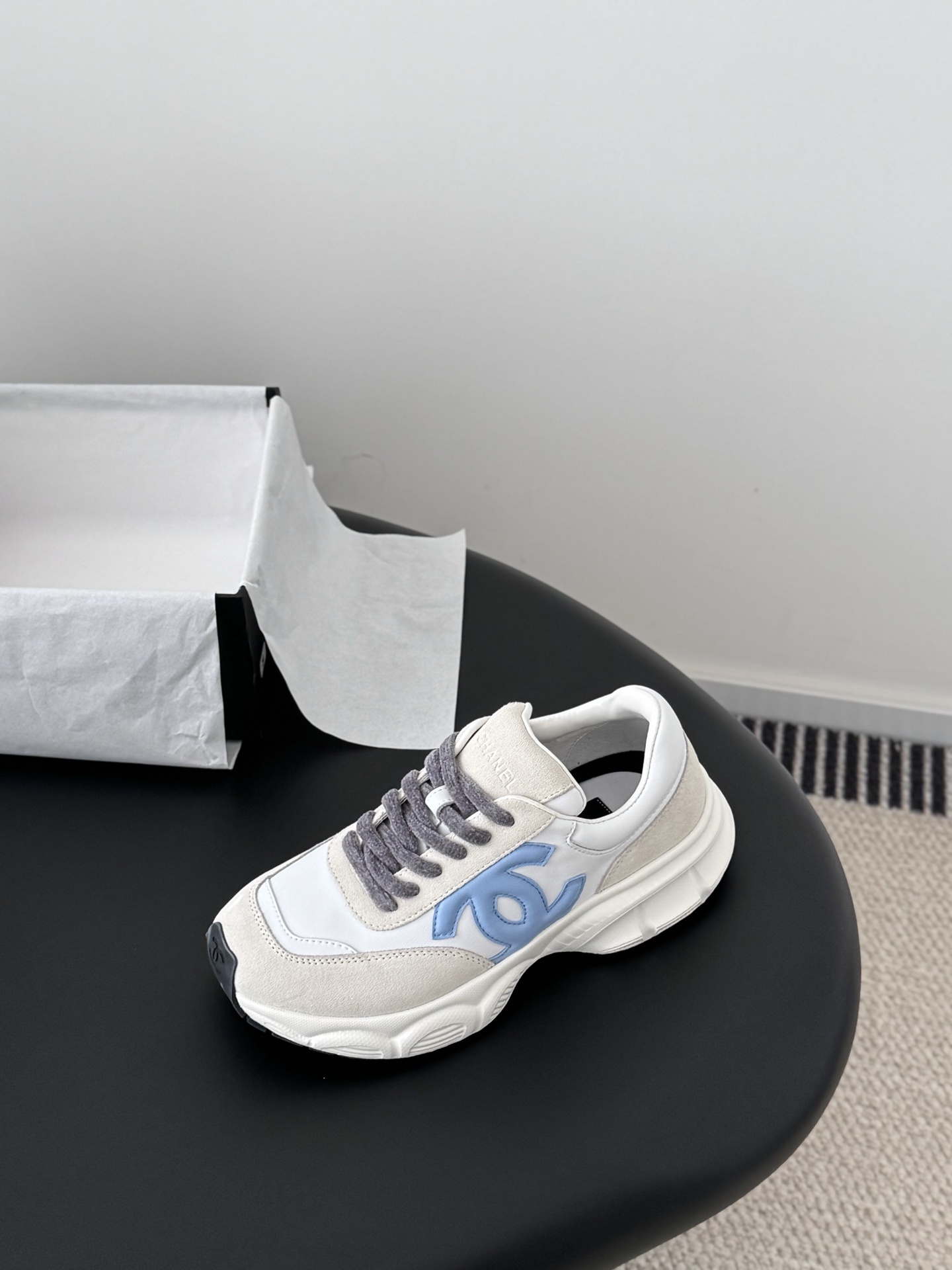 A pair of Chanel sneakers in white and beige suede with a light blue CC logo and grey laces, shown with a black Chanel shoebox. - Hình 6