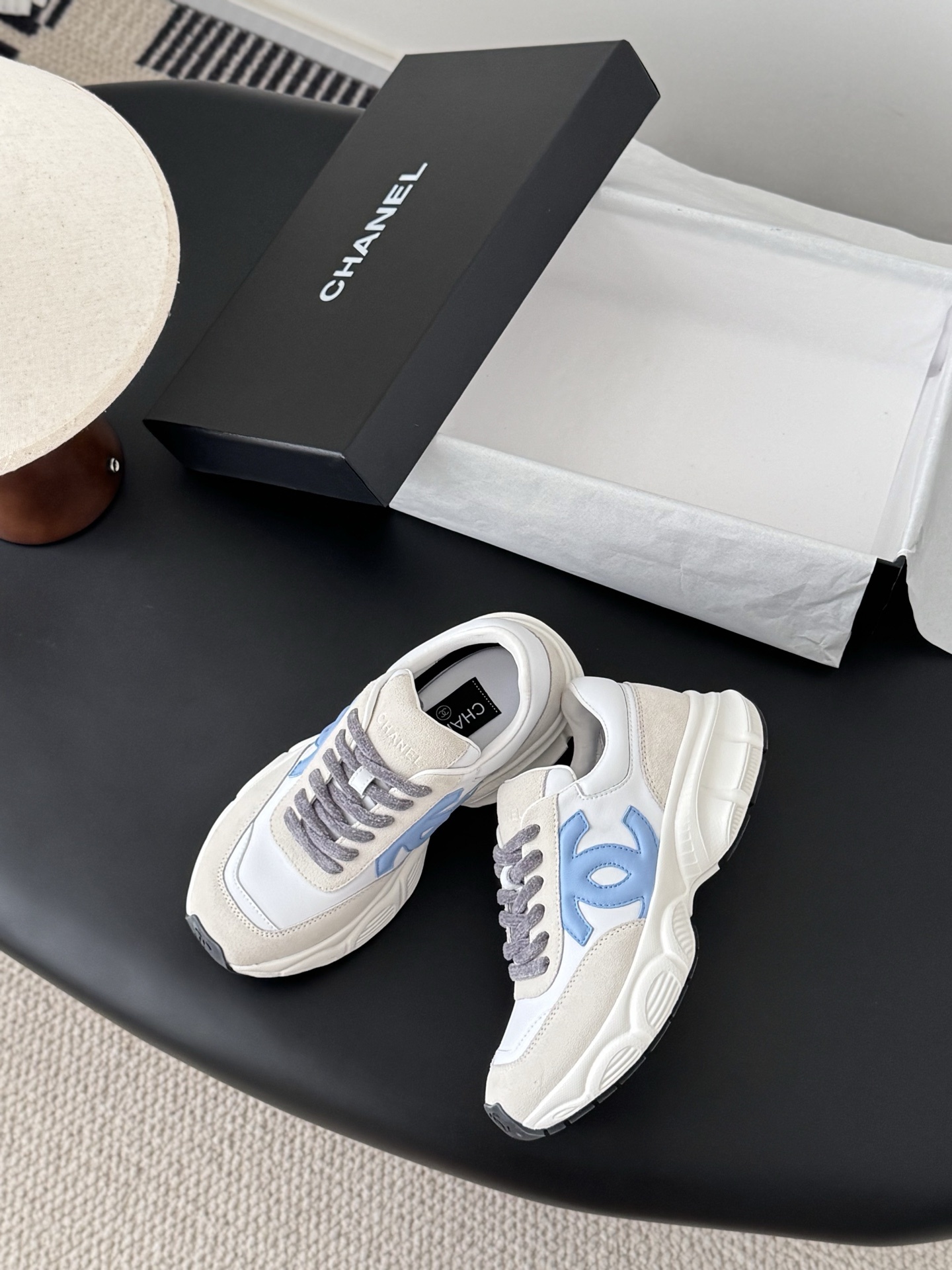 A pair of Chanel sneakers in white and beige suede with a light blue CC logo and grey laces, shown with a black Chanel shoebox. - Hình 3