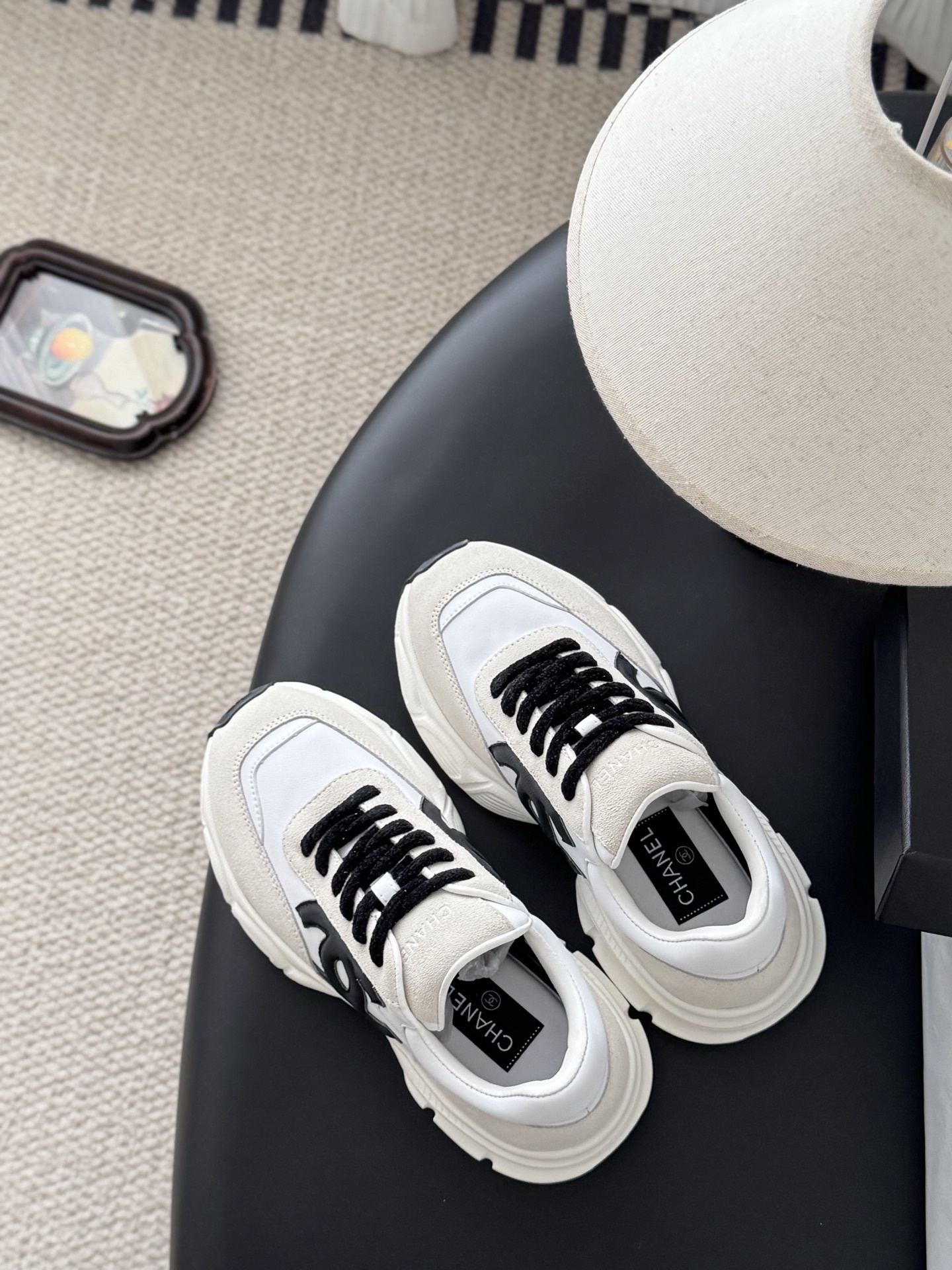 A pair of luxury Chanel white leather and beige suede sneakers with black CC logos and black laces, presented with a Chanel box on a black surface. - Hình 8