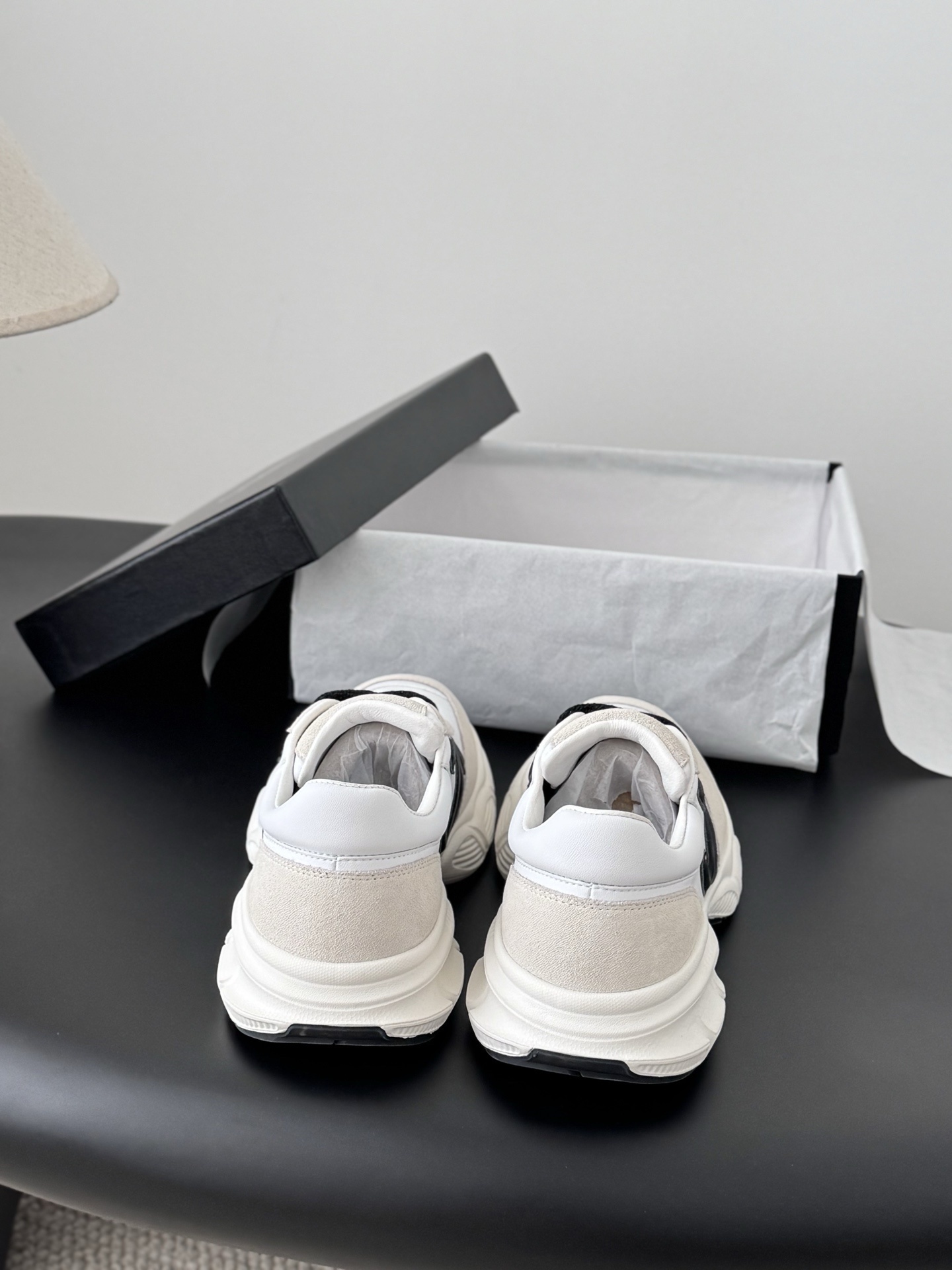 A pair of luxury Chanel white leather and beige suede sneakers with black CC logos and black laces, presented with a Chanel box on a black surface. - Hình 7