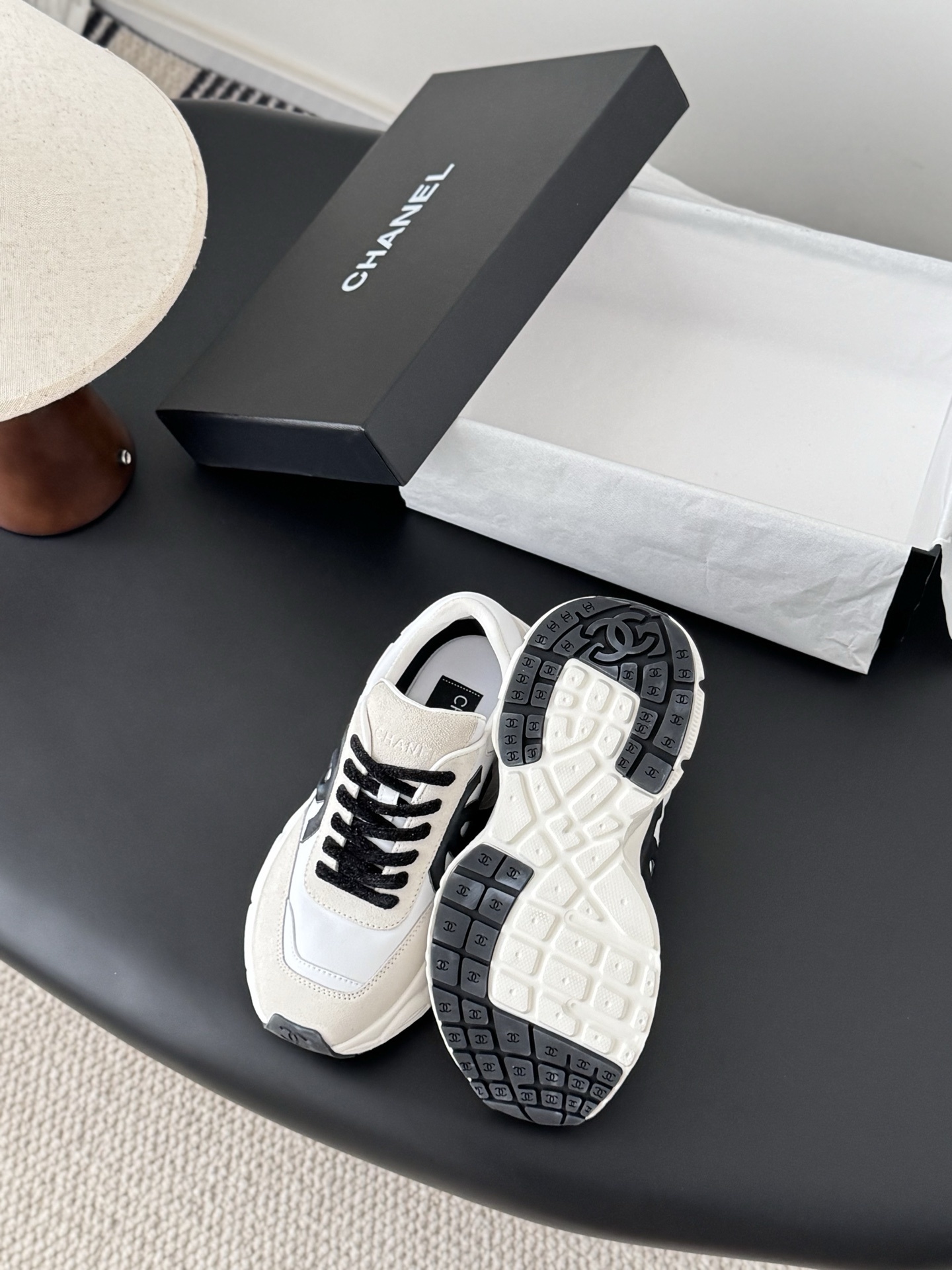 A pair of luxury Chanel white leather and beige suede sneakers with black CC logos and black laces, presented with a Chanel box on a black surface. - Hình 4