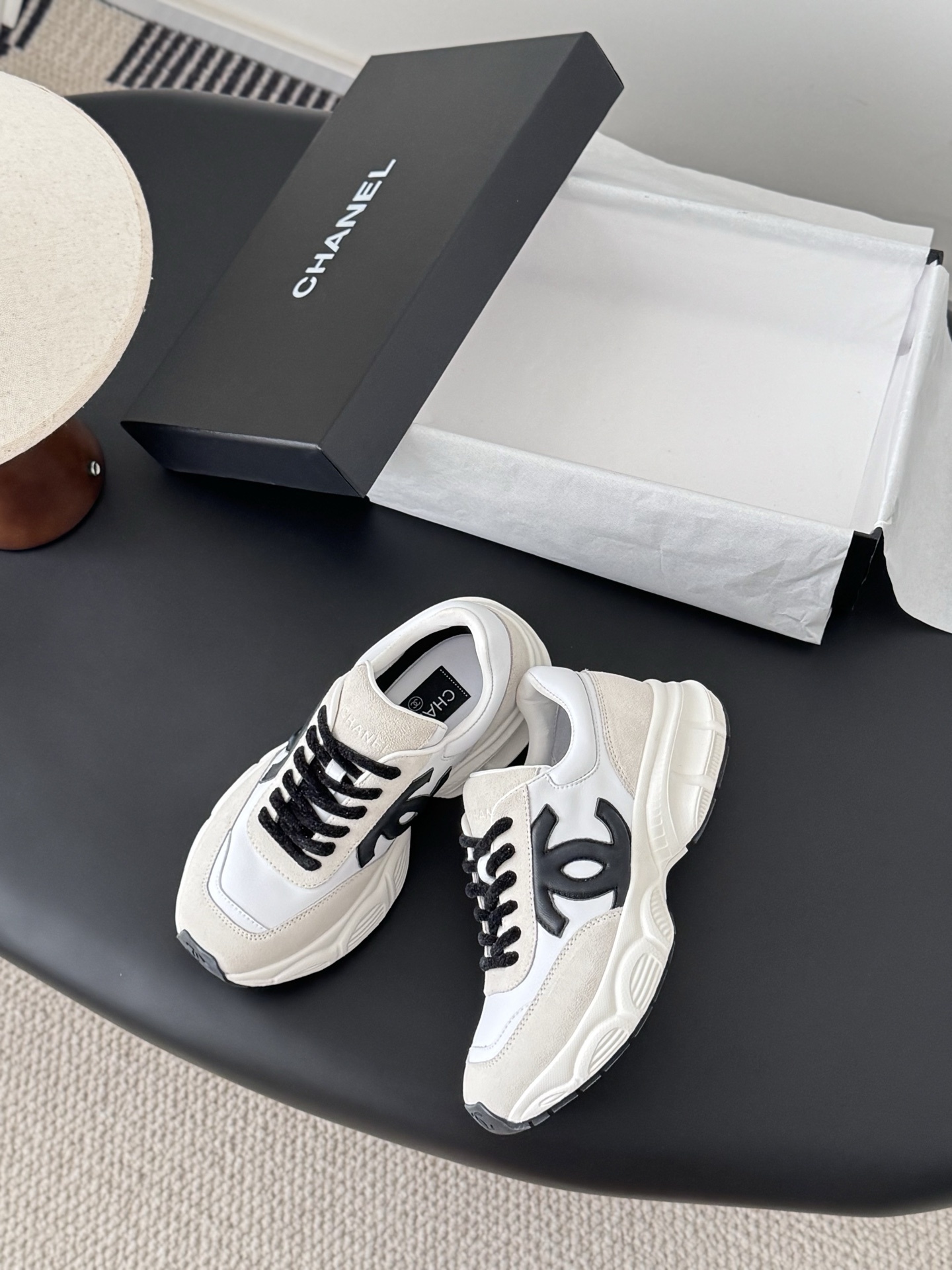 A pair of luxury Chanel white leather and beige suede sneakers with black CC logos and black laces, presented with a Chanel box on a black surface. - Hình 3