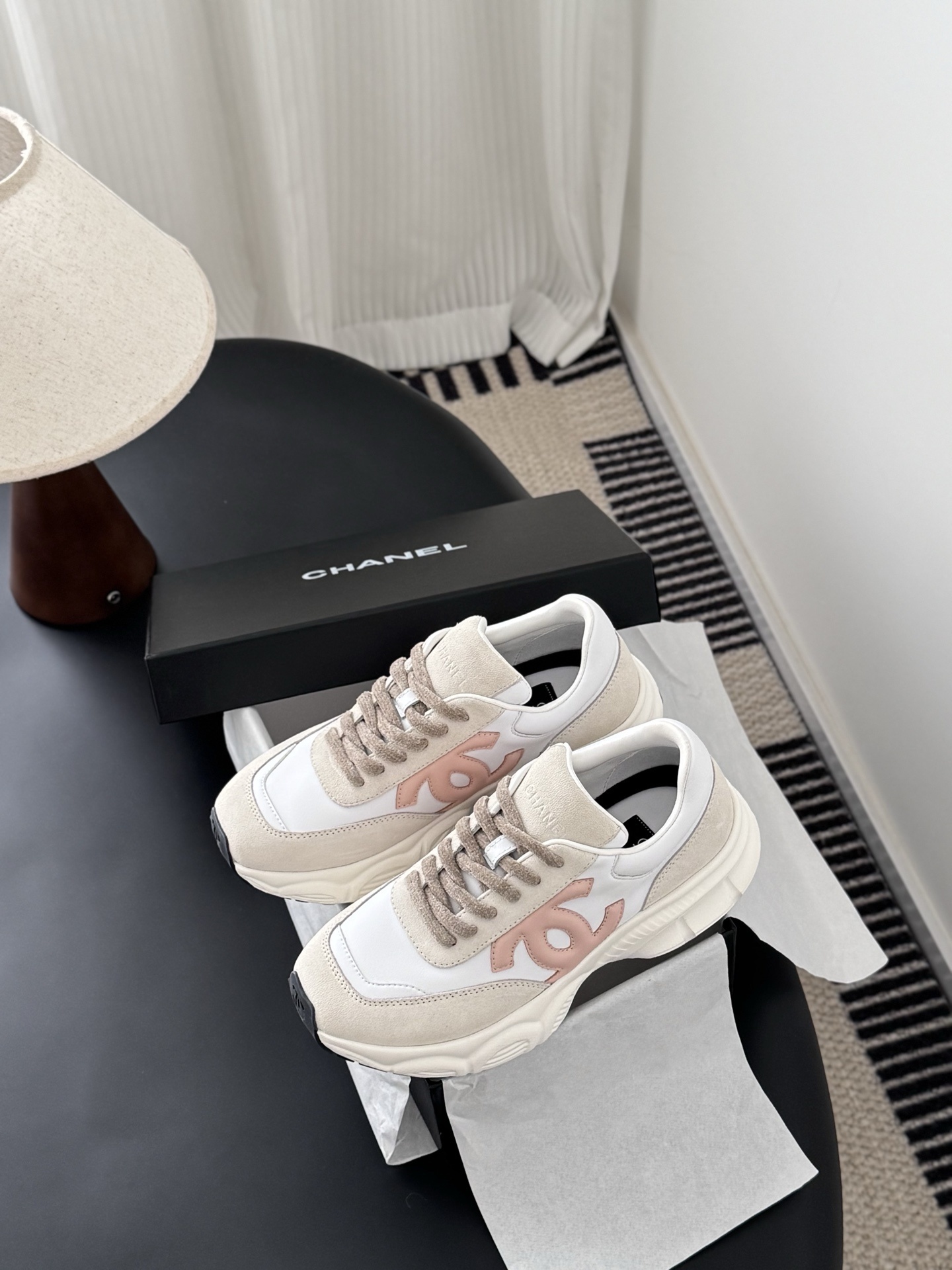 A pair of Chanel white and pink CC logo luxury sneakers displayed next to an open black Chanel shoebox on a dark table. - Hình 9