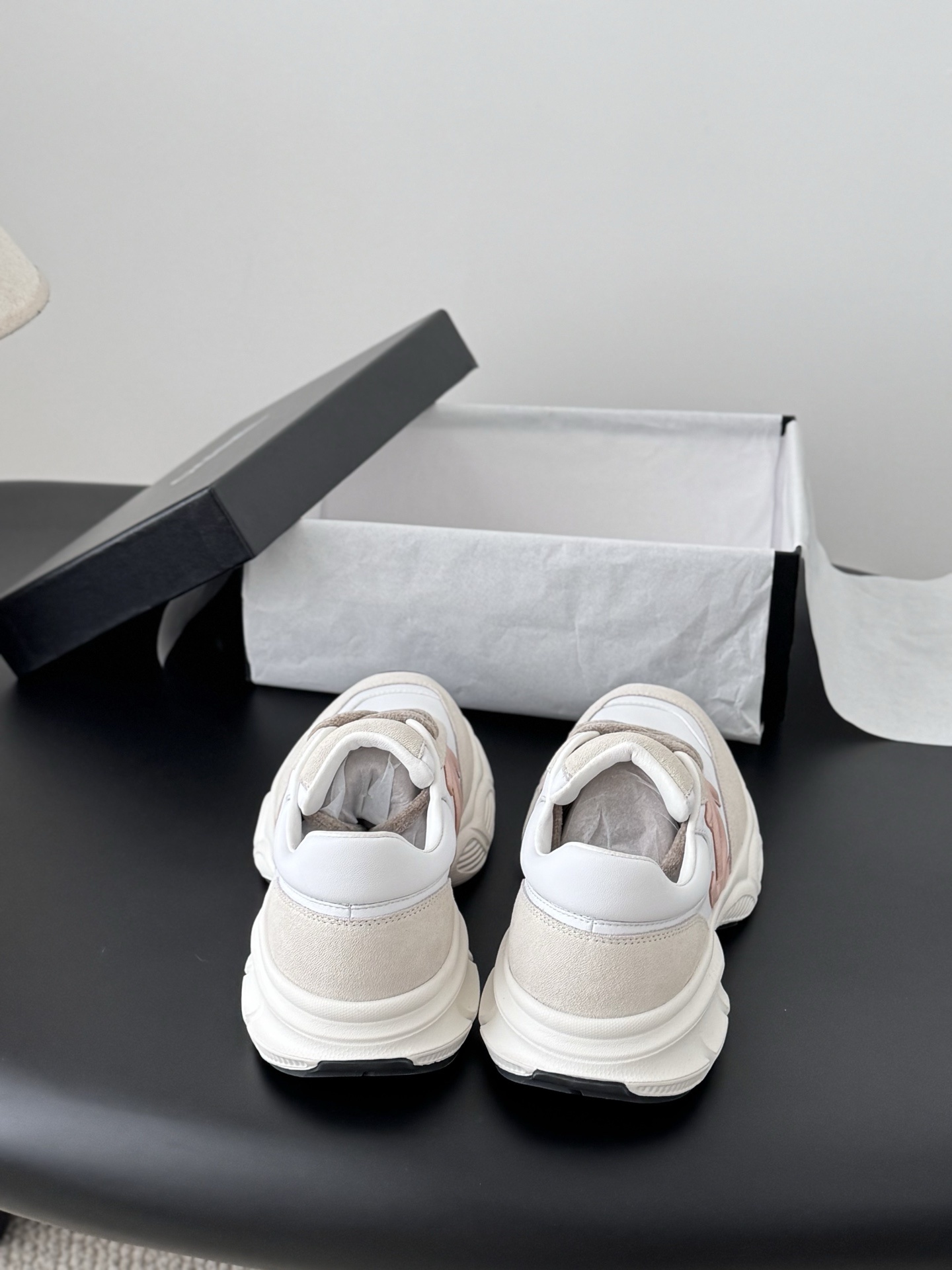 A pair of Chanel white and pink CC logo luxury sneakers displayed next to an open black Chanel shoebox on a dark table. - Hình 7