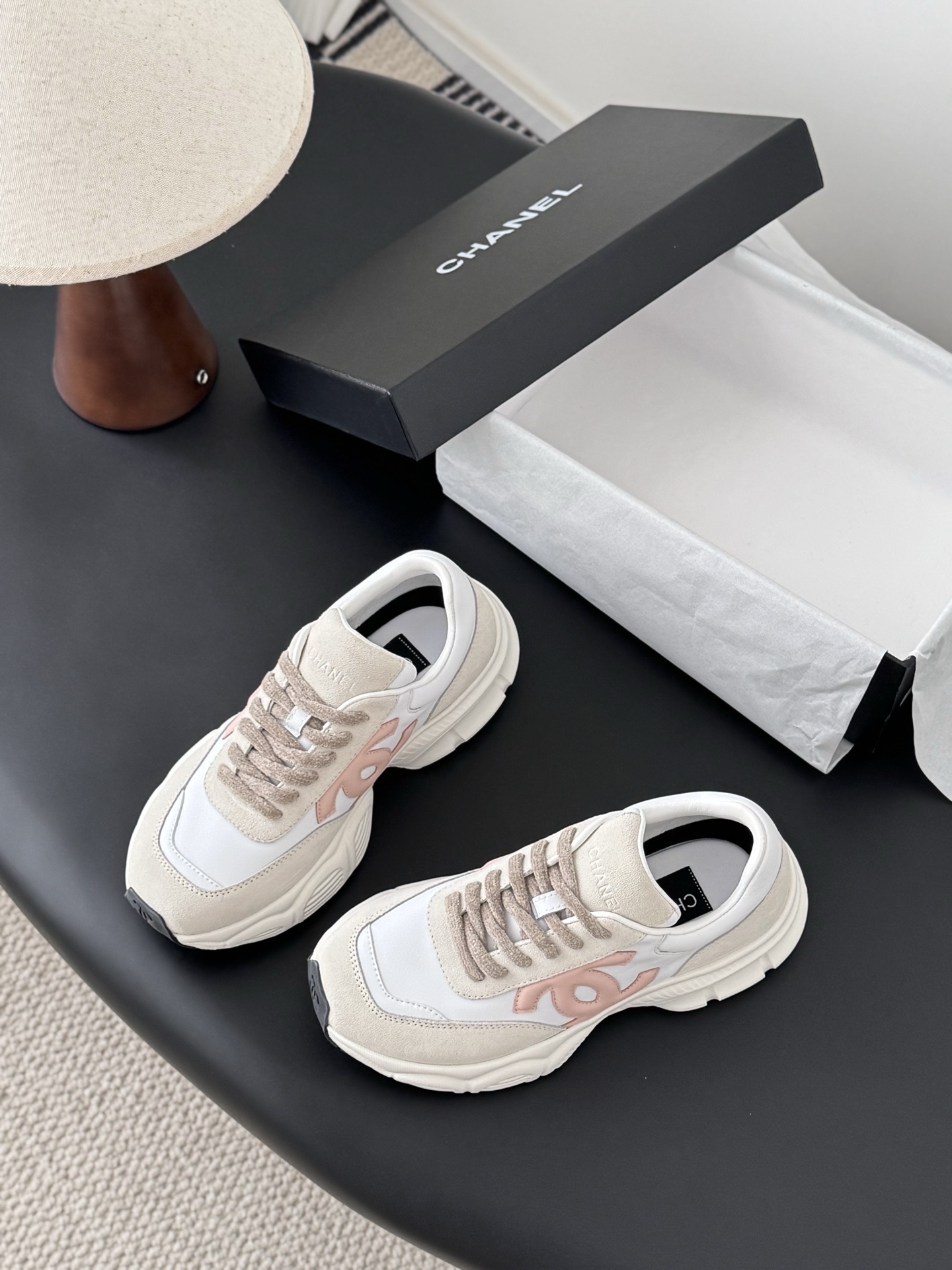 A pair of Chanel white and pink CC logo luxury sneakers displayed next to an open black Chanel shoebox on a dark table. - Hình 6