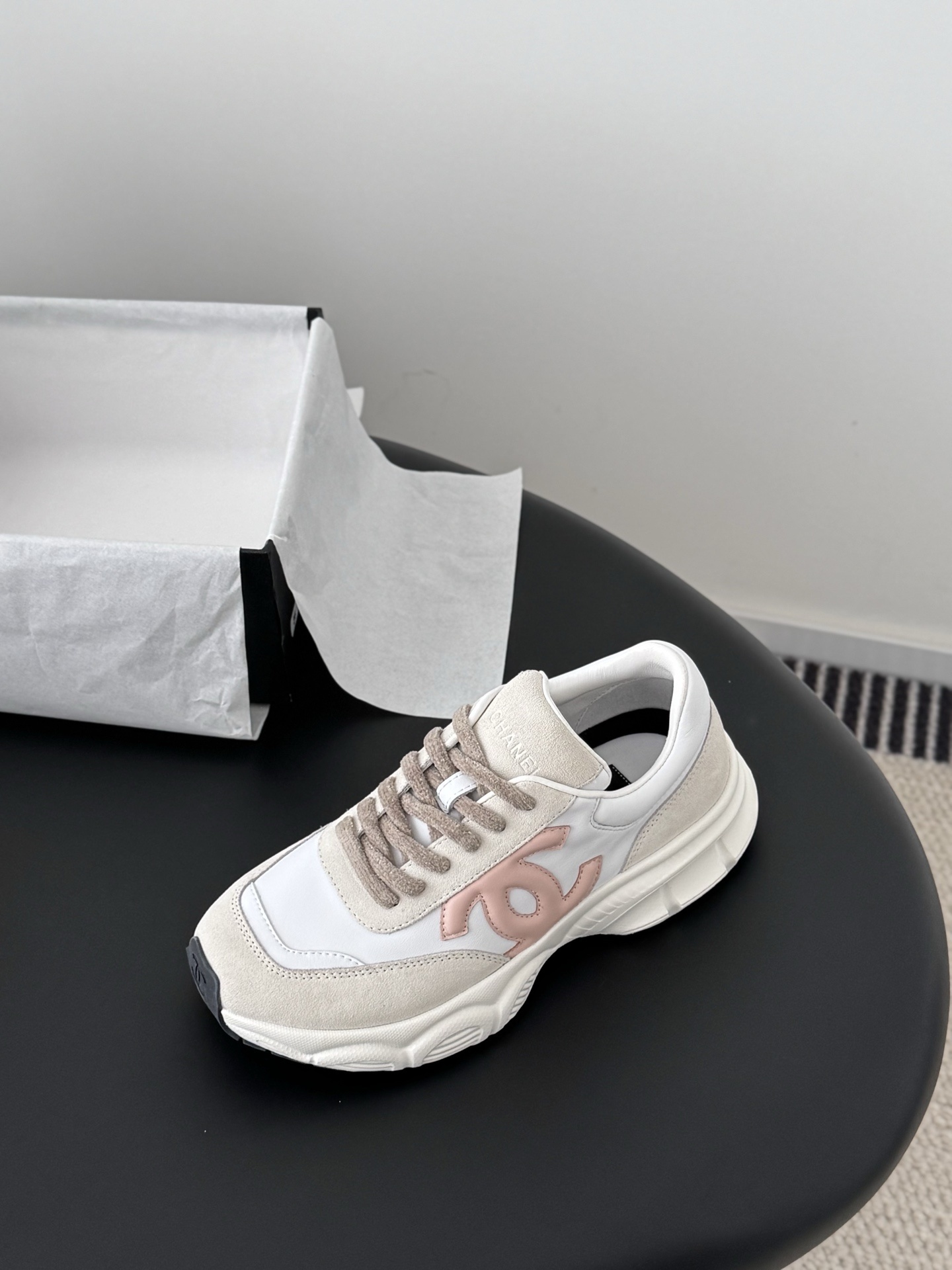 A pair of Chanel white and pink CC logo luxury sneakers displayed next to an open black Chanel shoebox on a dark table. - Hình 5