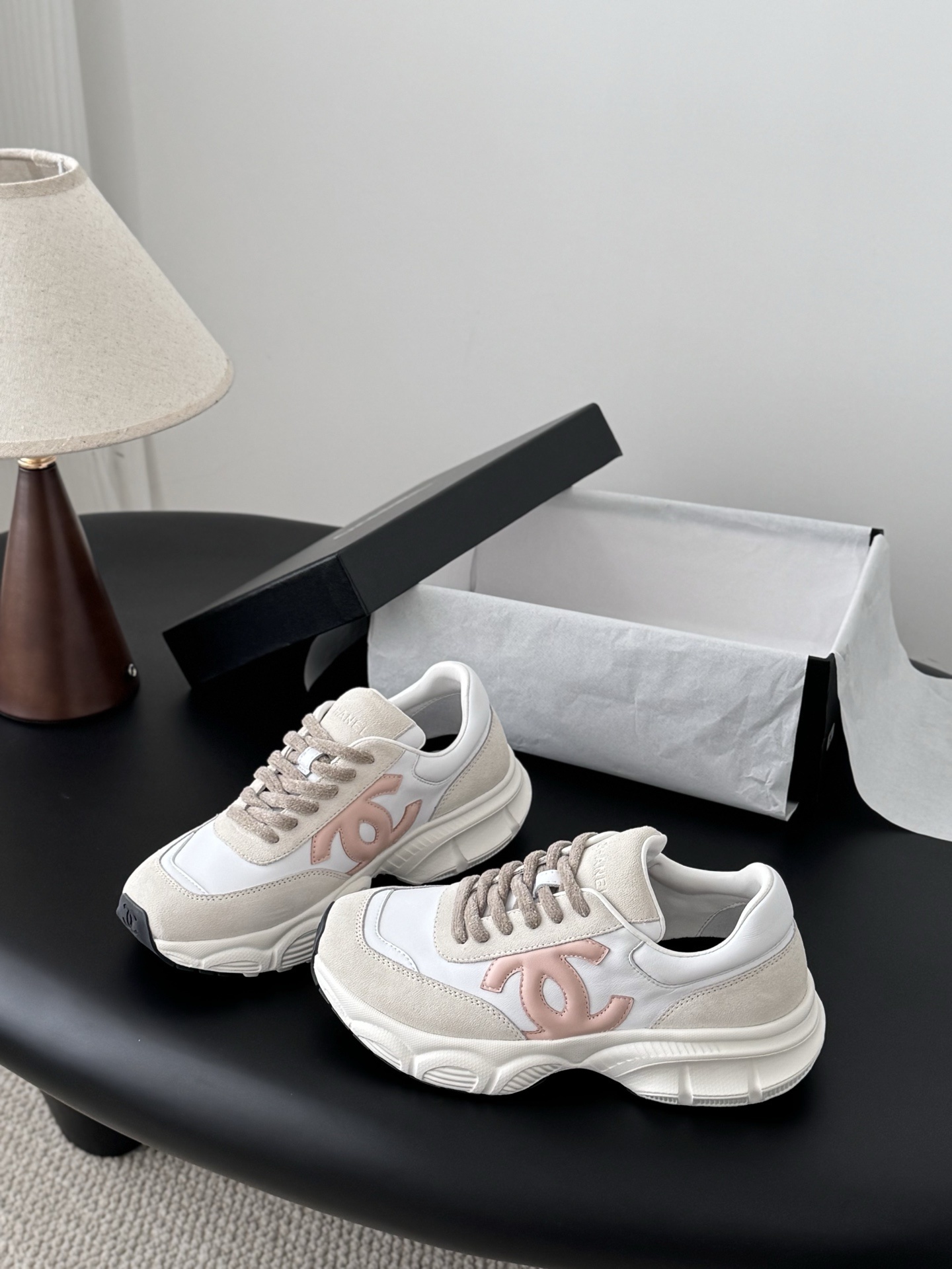 A pair of Chanel white and pink CC logo luxury sneakers displayed next to an open black Chanel shoebox on a dark table. - Hình 2