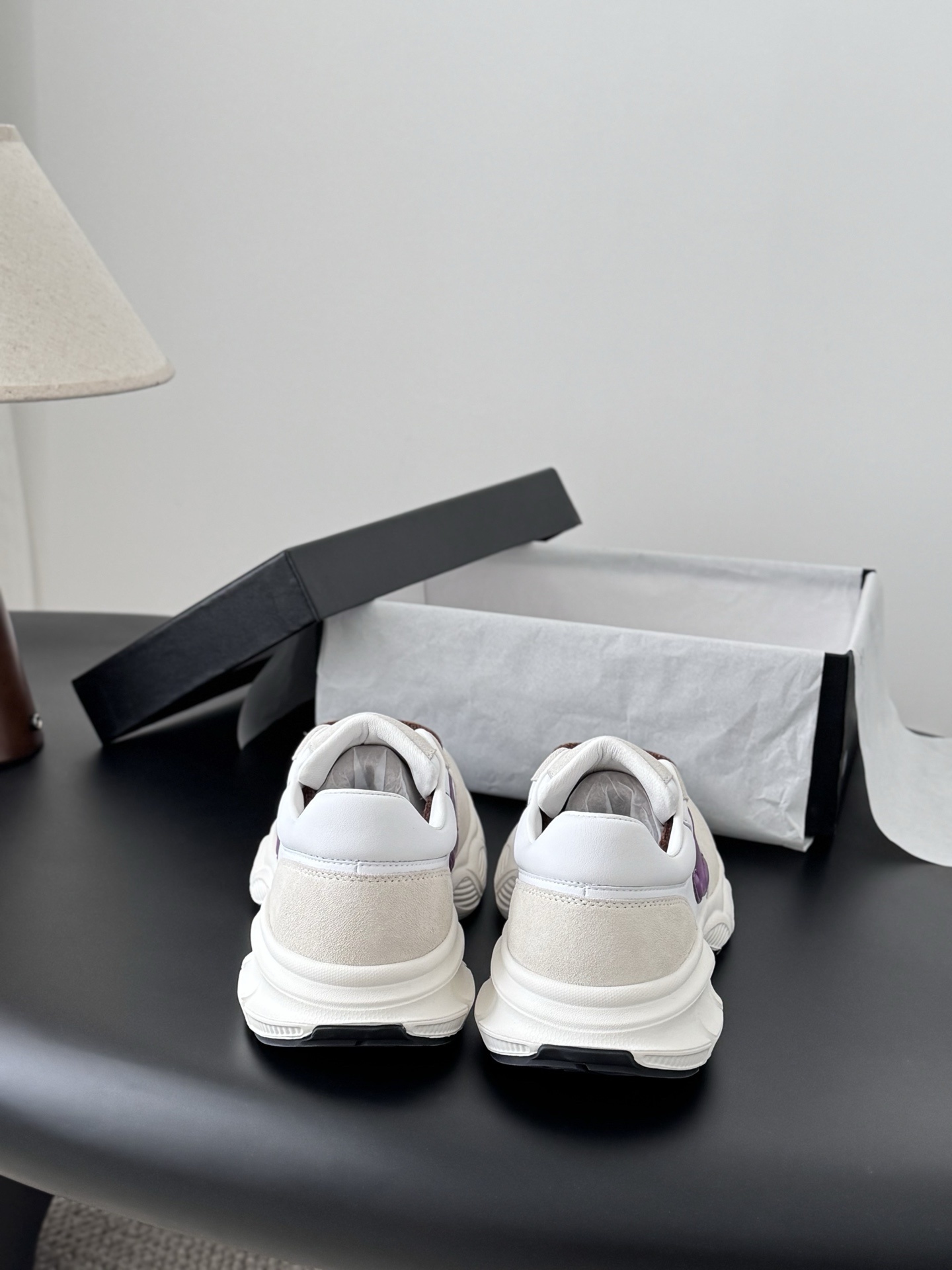 A pair of white Chanel sneakers with purple interlocking CC logos and brown laces next to a black Chanel box. - Hình 6