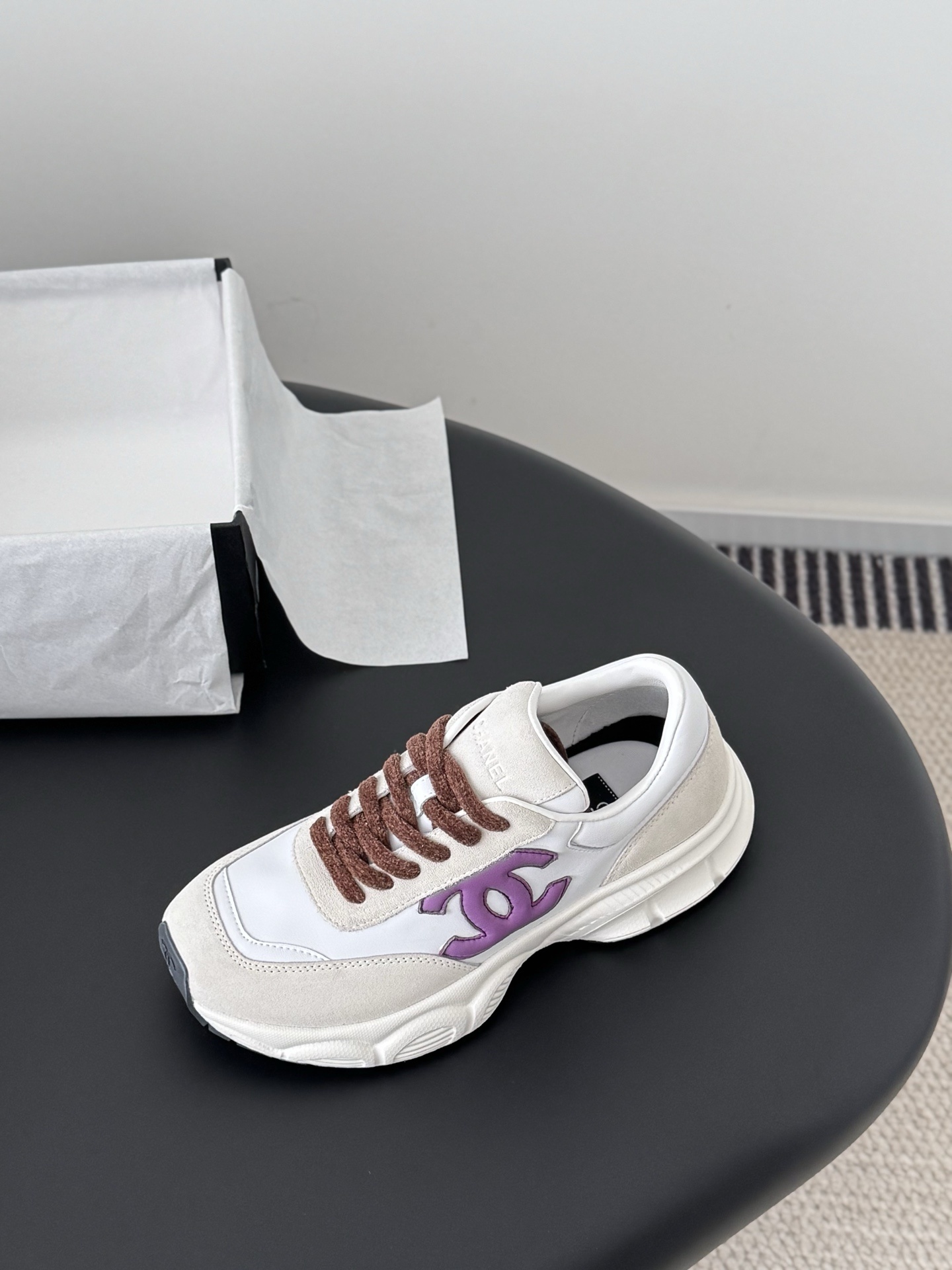 A pair of white Chanel sneakers with purple interlocking CC logos and brown laces next to a black Chanel box. - Hình 5