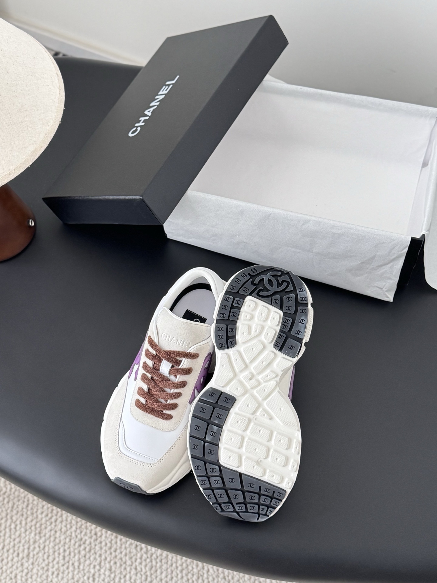 A pair of white Chanel sneakers with purple interlocking CC logos and brown laces next to a black Chanel box. - Hình 4