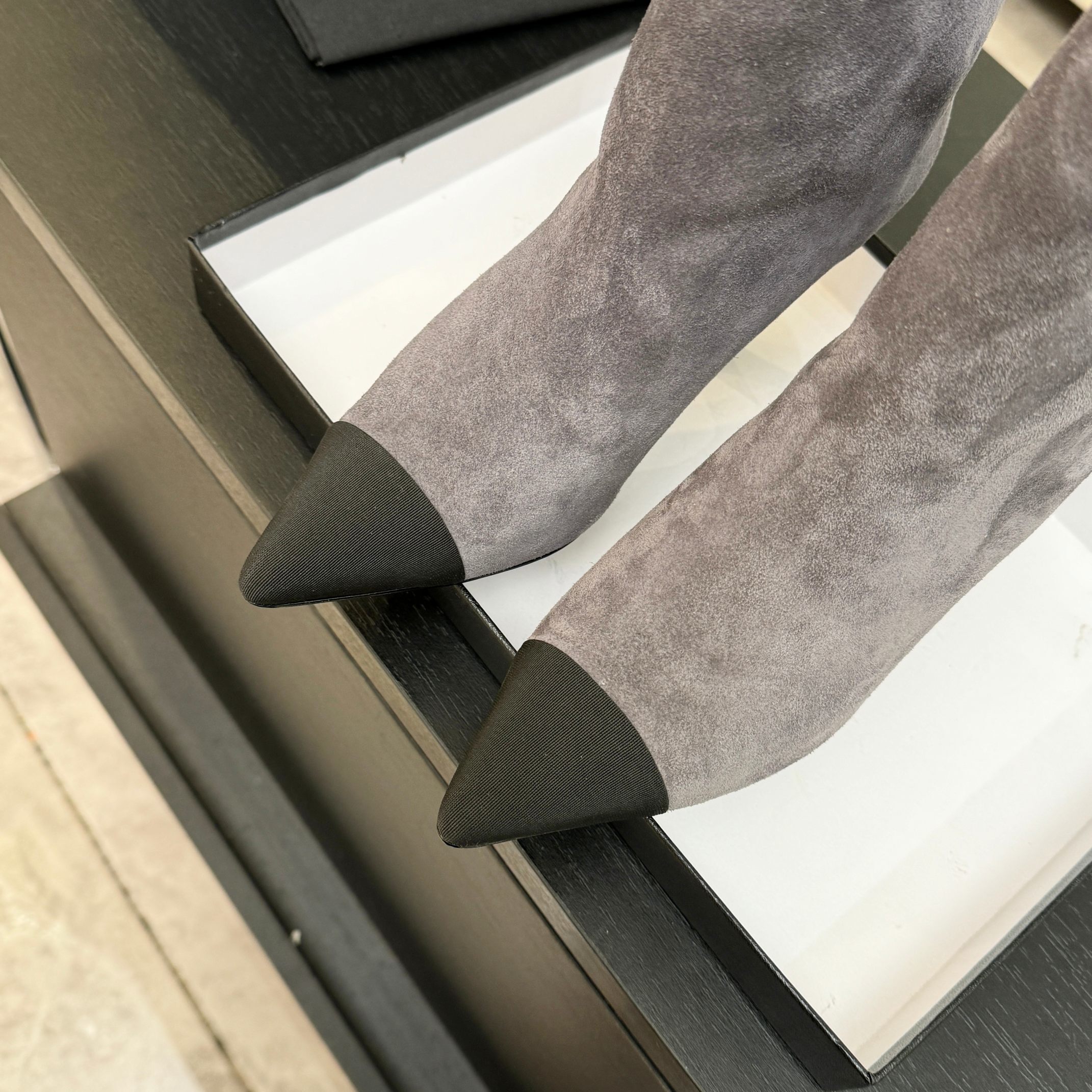 A pair of luxury gray Chanel suede ankle boots with black pointed cap toes and Gabrielle Chanel embroidery displayed on a black surface. - Hình 9