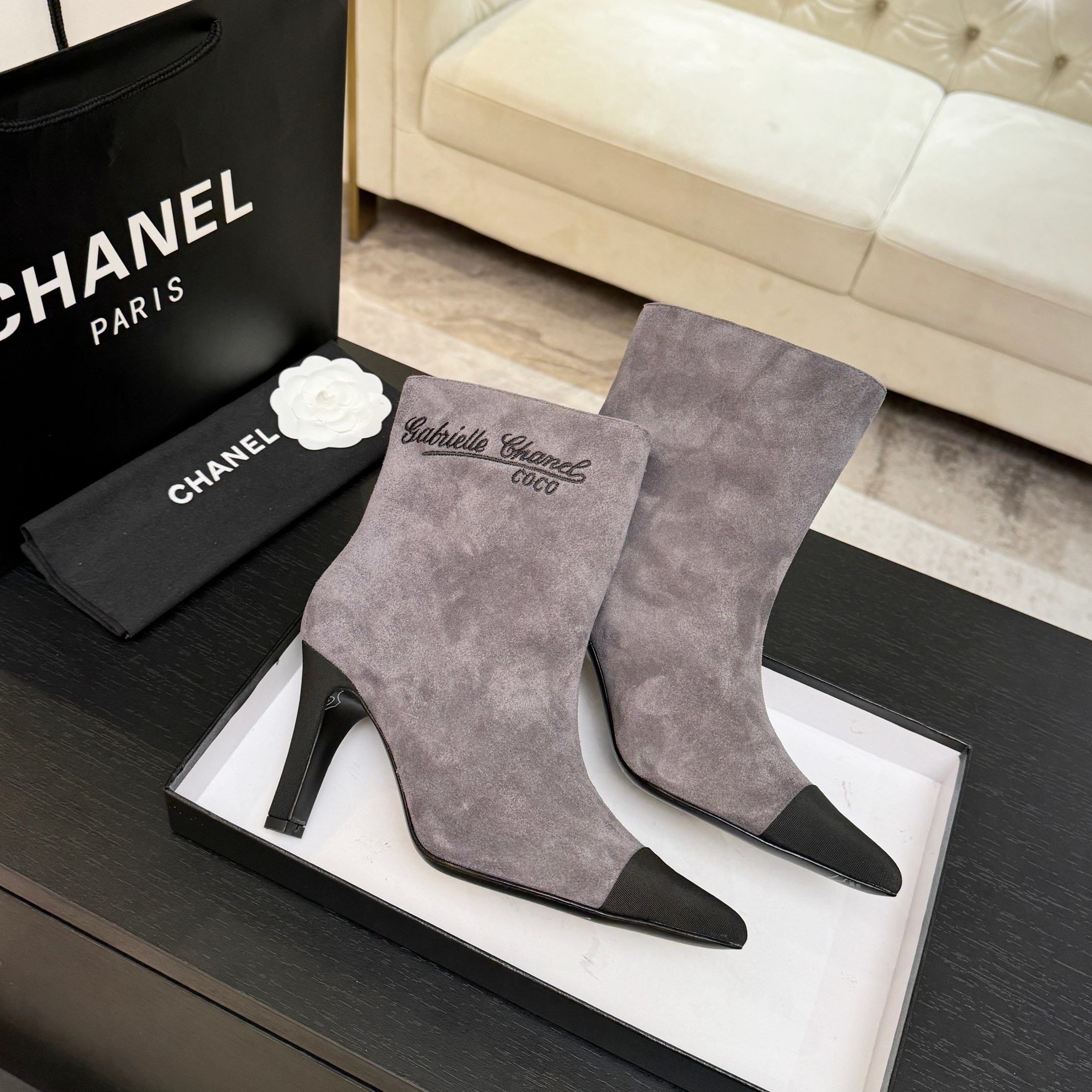 A pair of luxury gray Chanel suede ankle boots with black pointed cap toes and Gabrielle Chanel embroidery displayed on a black surface. - Hình 7