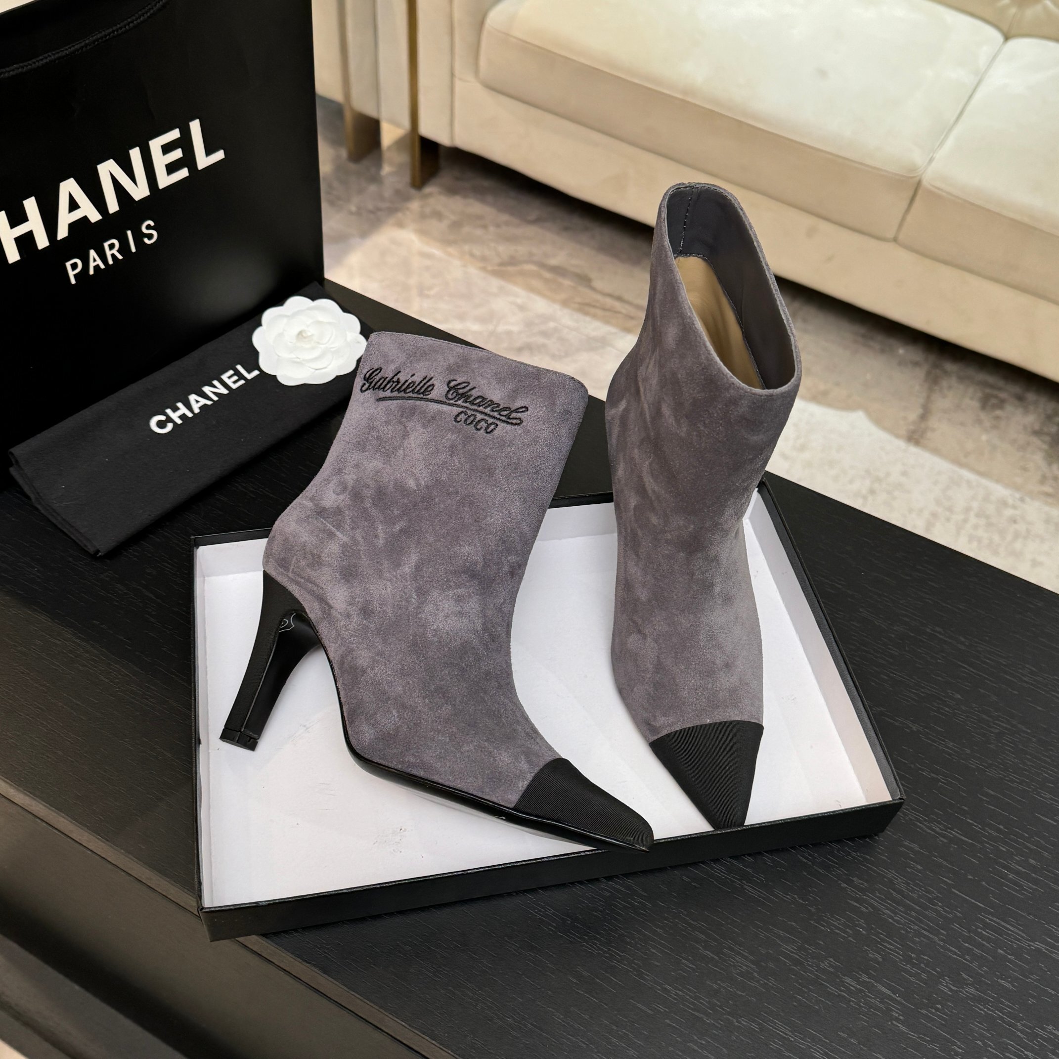 A pair of luxury gray Chanel suede ankle boots with black pointed cap toes and Gabrielle Chanel embroidery displayed on a black surface. - Hình 6