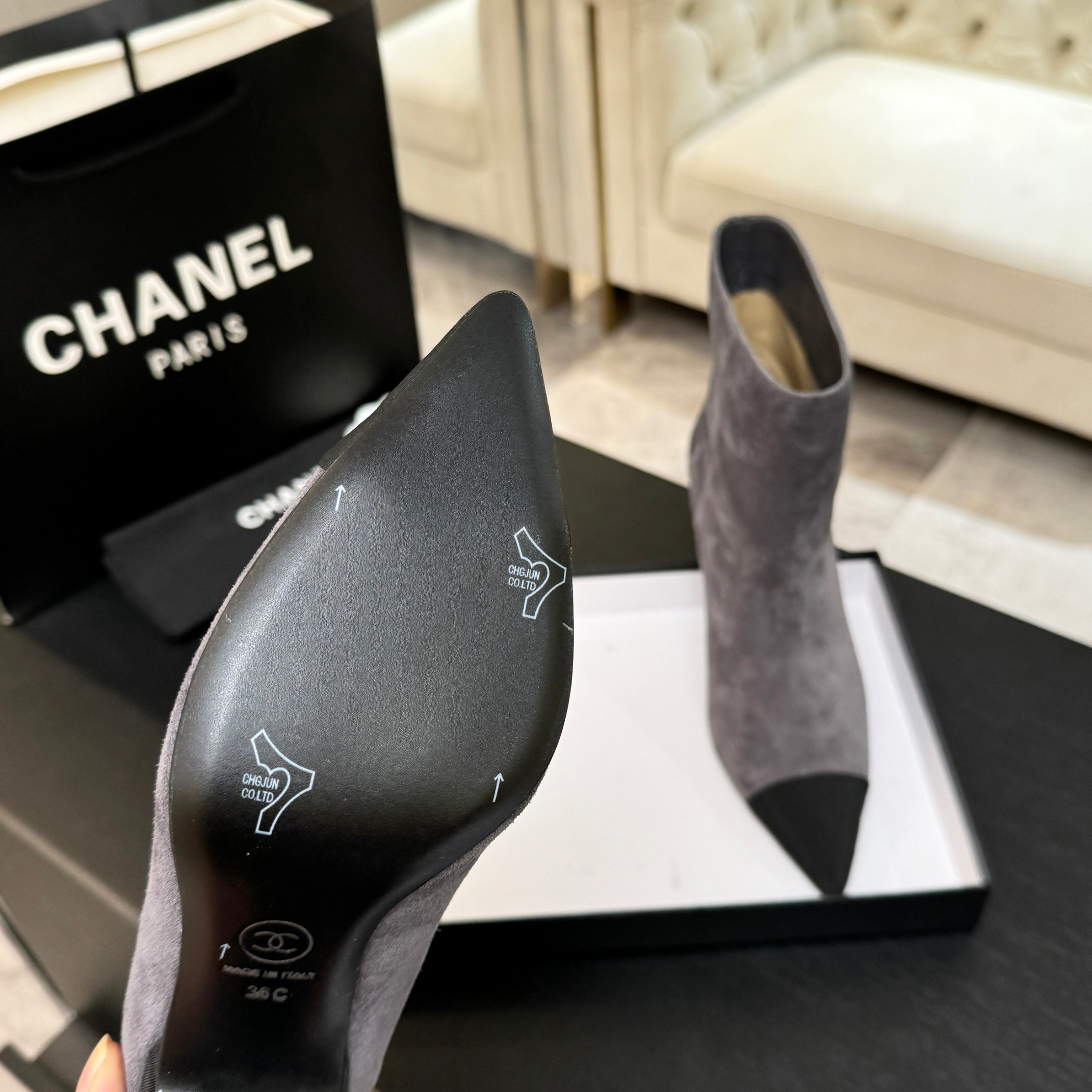 A pair of luxury gray Chanel suede ankle boots with black pointed cap toes and Gabrielle Chanel embroidery displayed on a black surface. - Hình 5