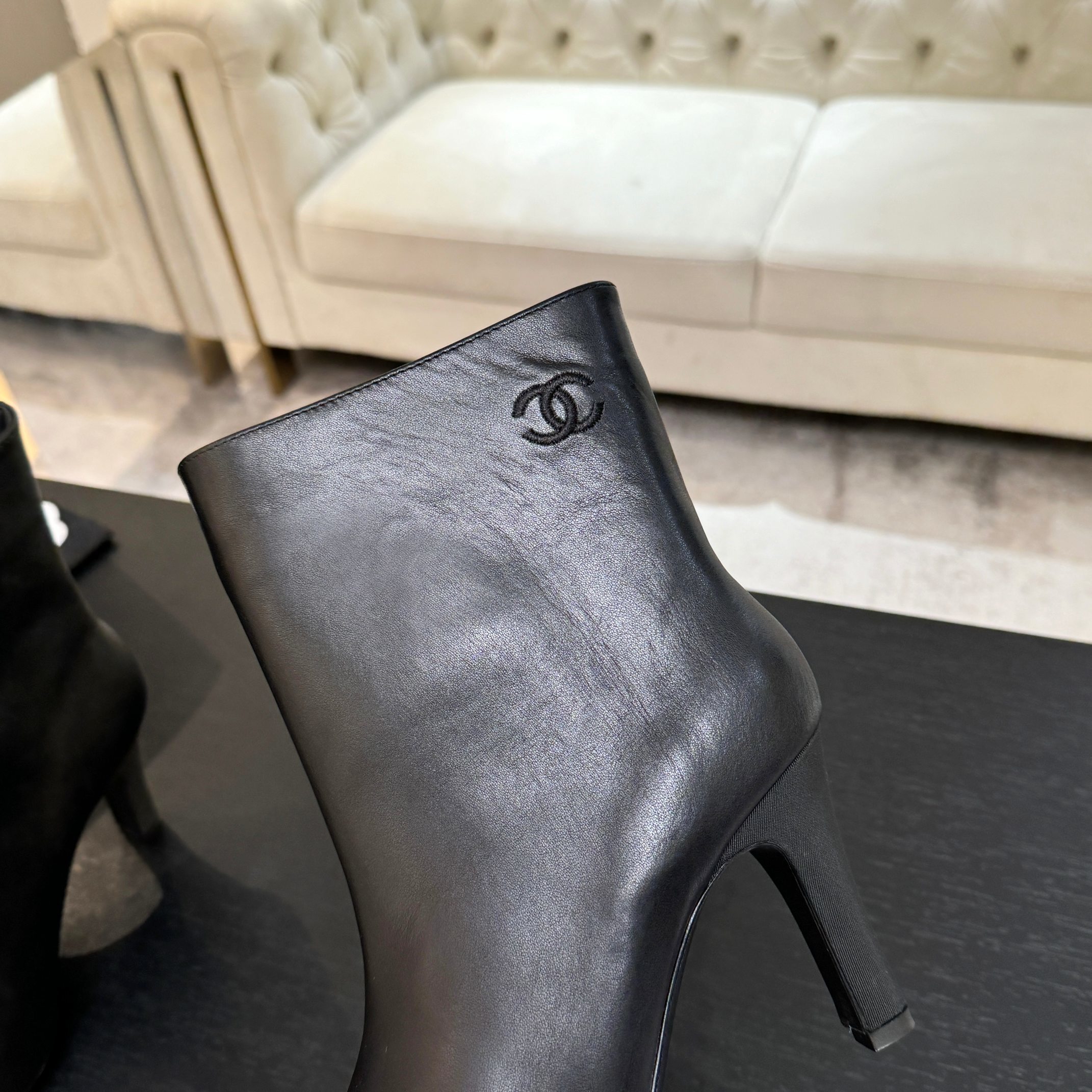 Pair of black Chanel Gabrielle leather ankle boots with pointed toes and ribbed fabric cap toes displayed on a black surface next to a Chanel shopping bag. - Hình 9