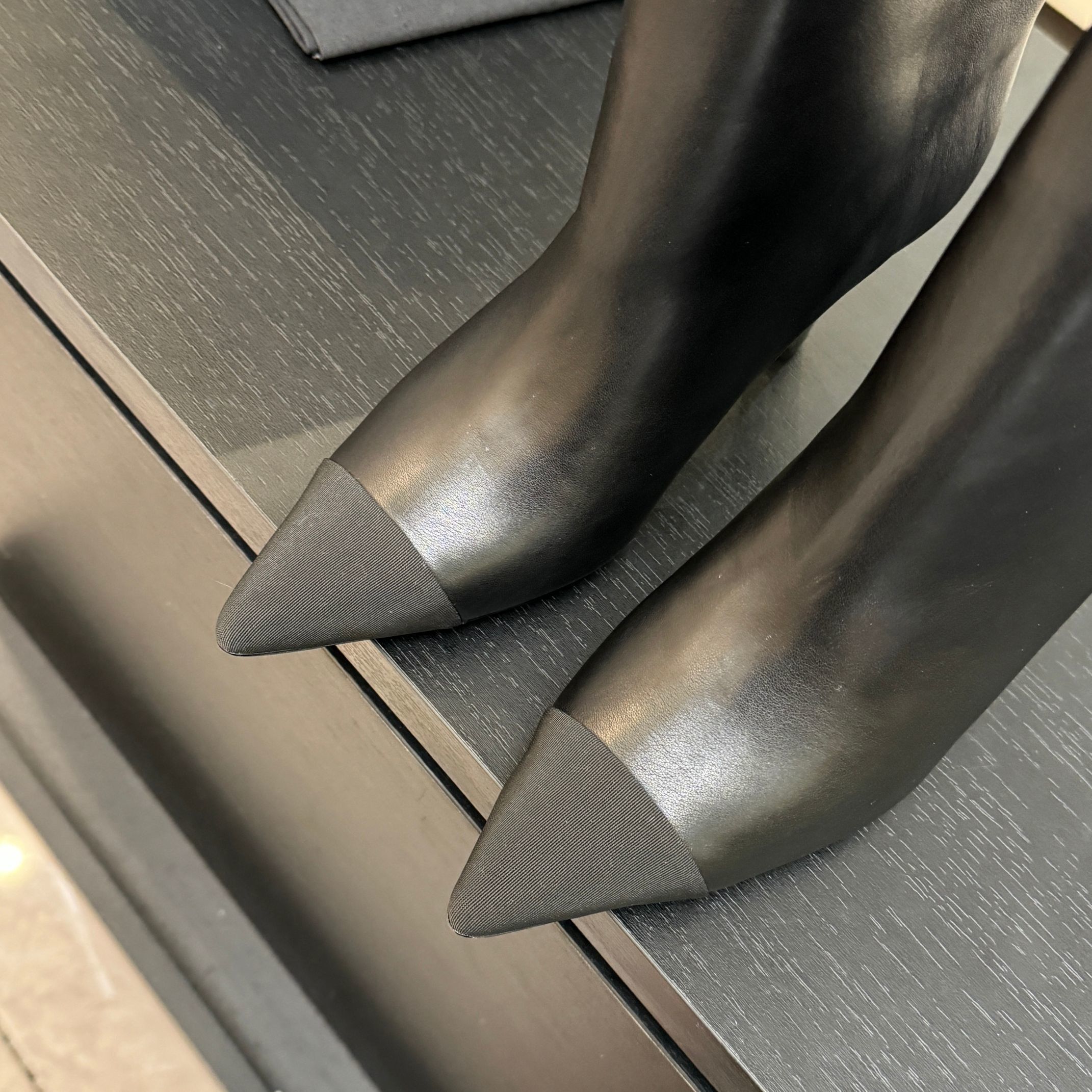 Pair of black Chanel Gabrielle leather ankle boots with pointed toes and ribbed fabric cap toes displayed on a black surface next to a Chanel shopping bag. - Hình 8