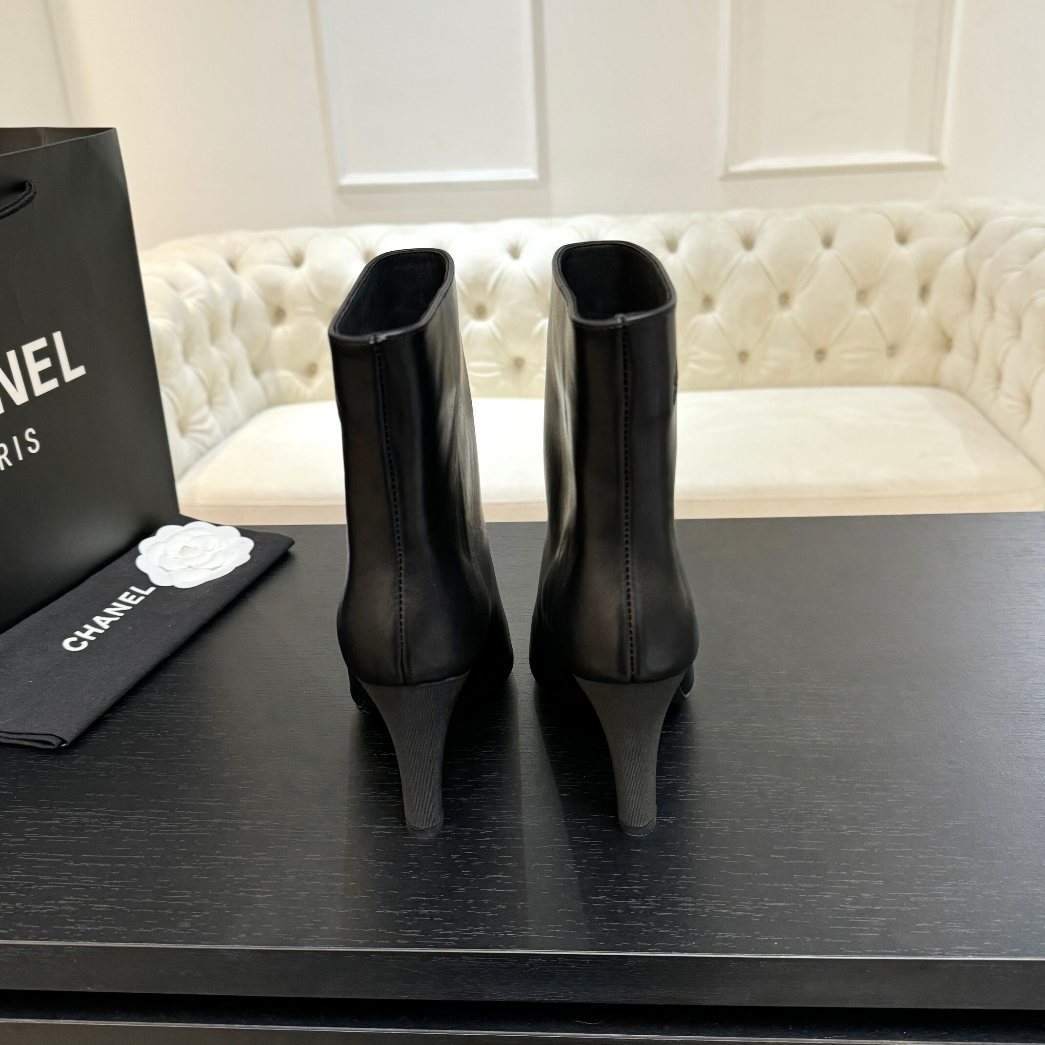 Pair of black Chanel Gabrielle leather ankle boots with pointed toes and ribbed fabric cap toes displayed on a black surface next to a Chanel shopping bag. - Hình 5