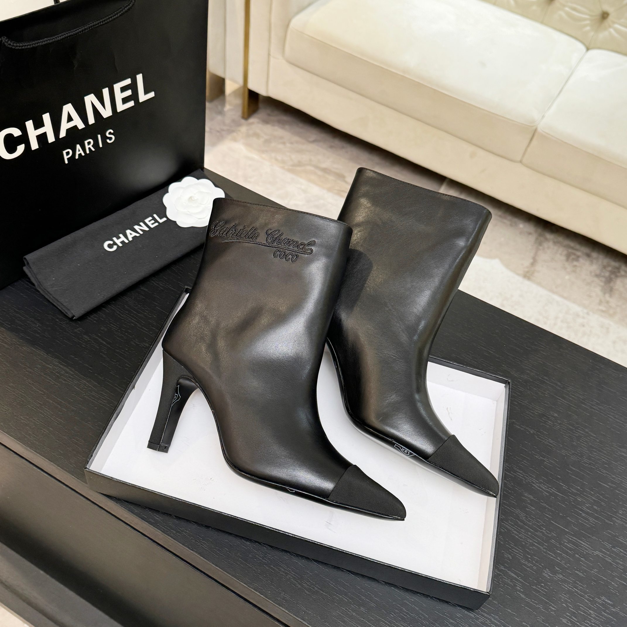 Pair of black Chanel Gabrielle leather ankle boots with pointed toes and ribbed fabric cap toes displayed on a black surface next to a Chanel shopping bag. - Hình 4