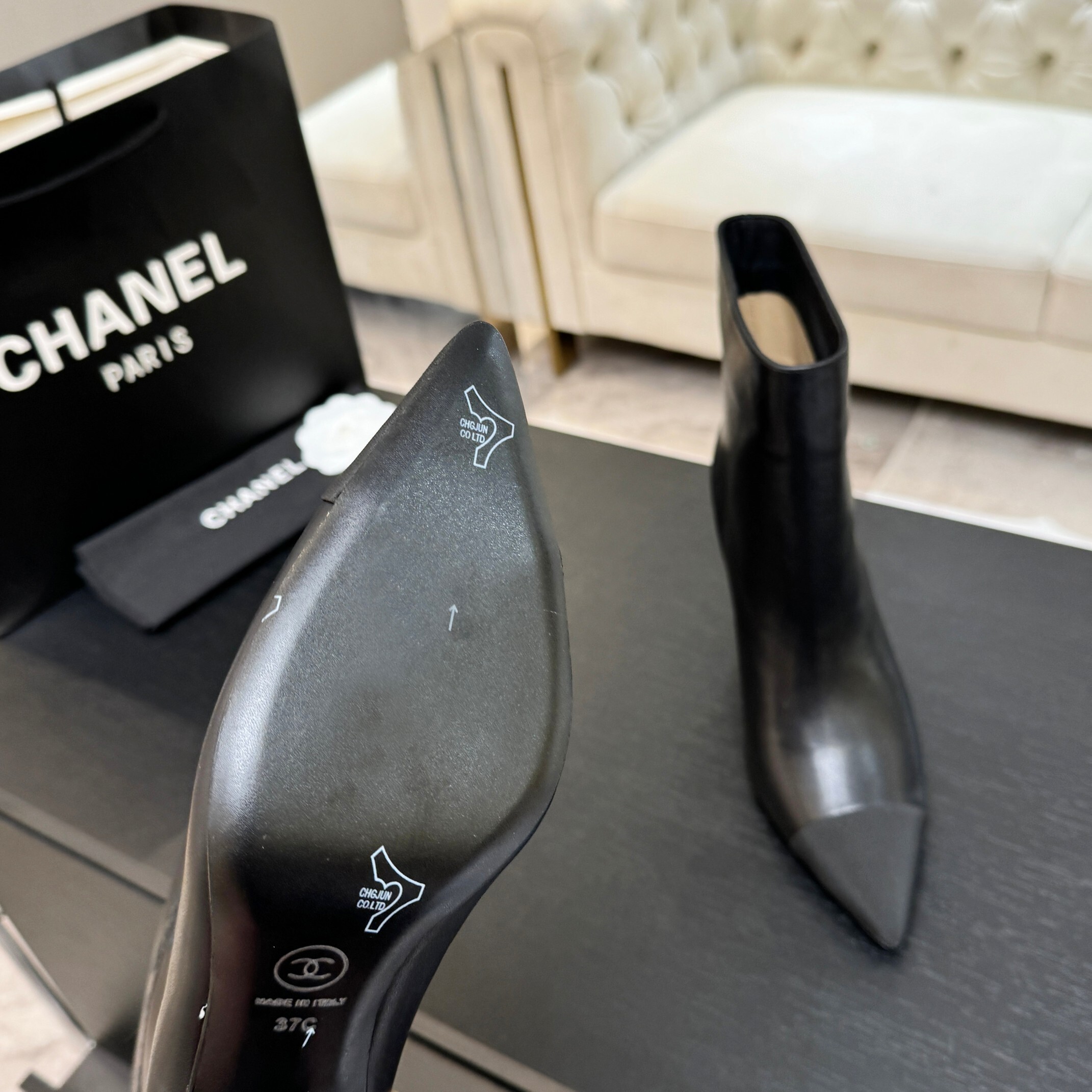 Pair of black Chanel Gabrielle leather ankle boots with pointed toes and ribbed fabric cap toes displayed on a black surface next to a Chanel shopping bag. - Hình 3