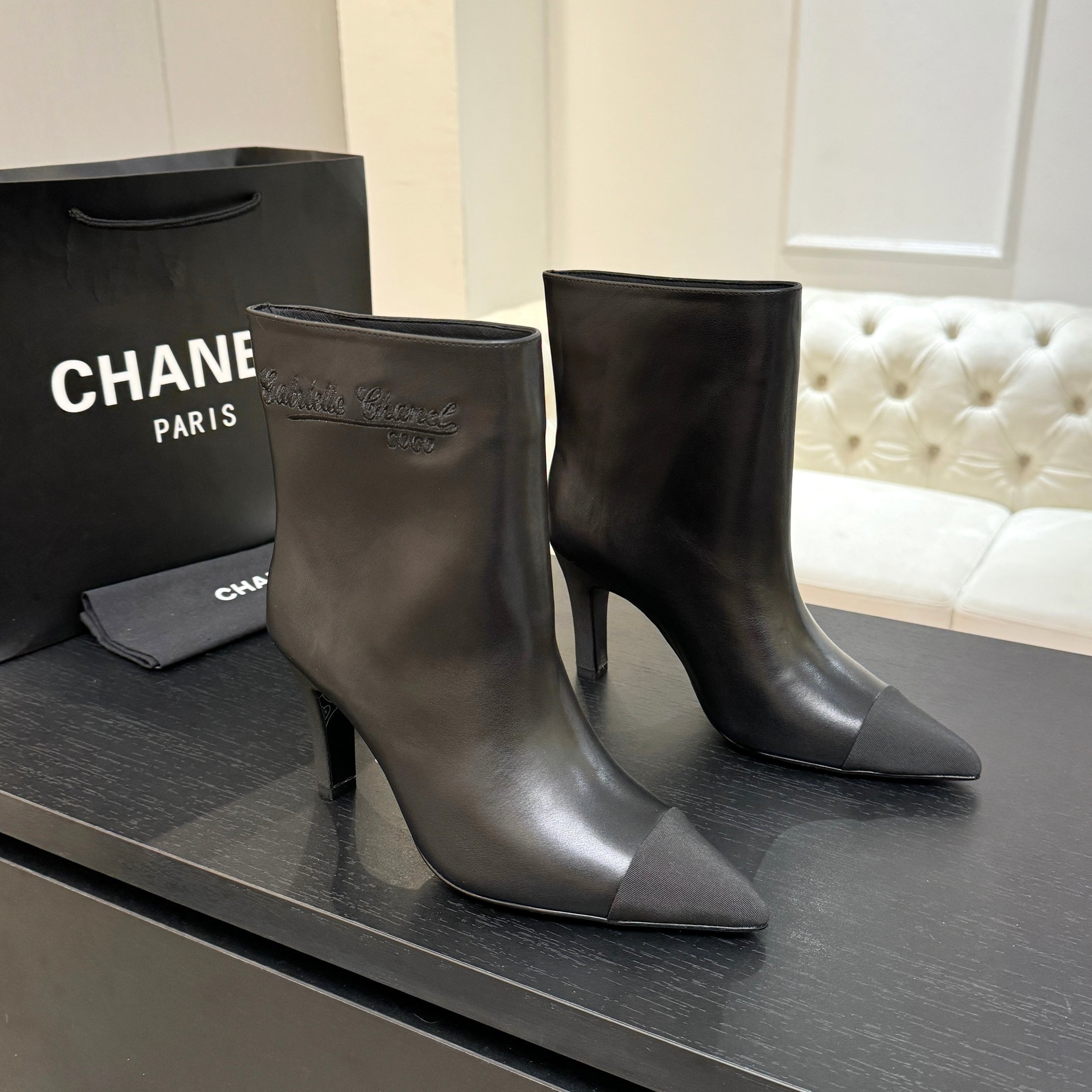 Pair of black Chanel Gabrielle leather ankle boots with pointed toes and ribbed fabric cap toes displayed on a black surface next to a Chanel shopping bag. - Hình 2