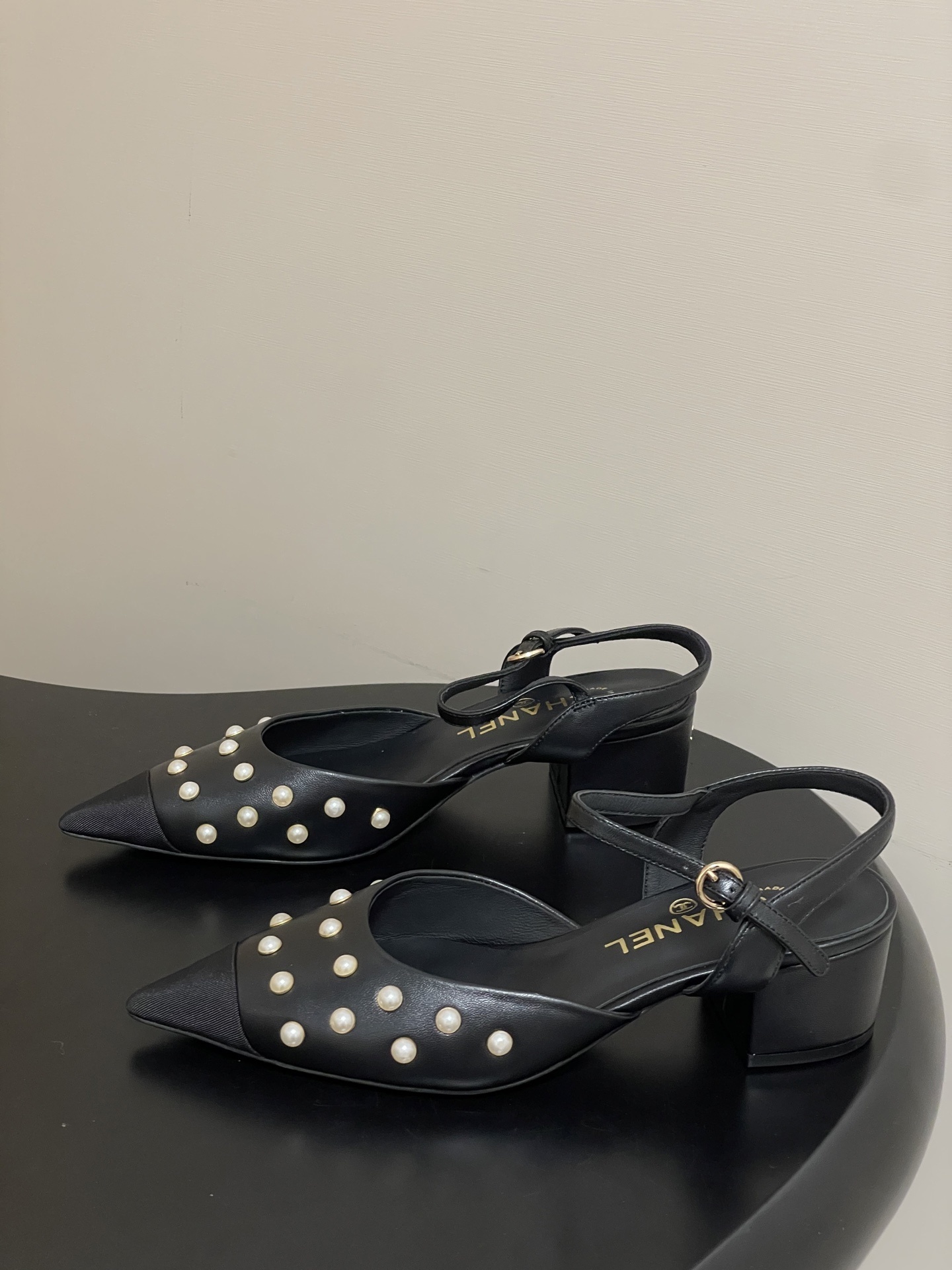 A pair of Chanel black leather slingback pumps with pearl studs and block heels displayed on a black surface. - Hình 3