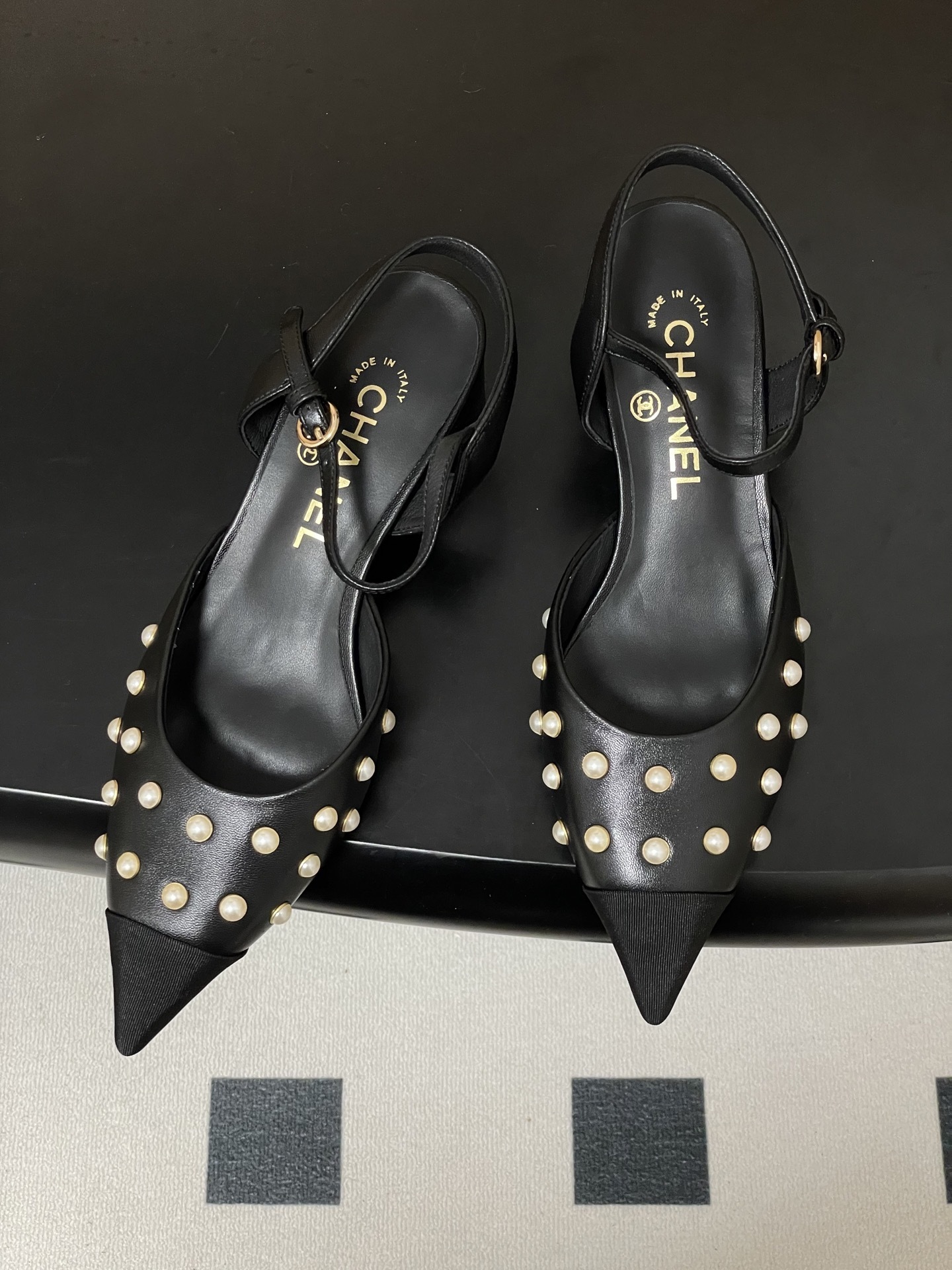 A pair of Chanel black leather slingback pumps with pearl studs and block heels displayed on a black surface. - Hình 2