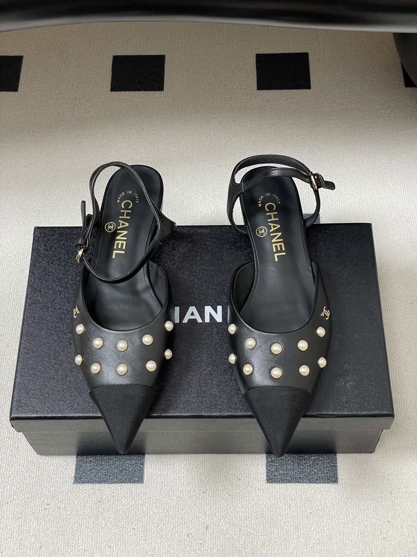 A pair of black Chanel slingback flats with pearl studs, pointed fabric cap toes, and gold CC logo on a black surface. - Hình 5