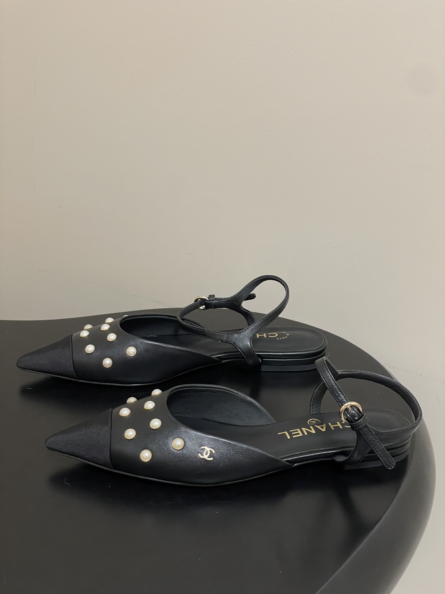 A pair of black Chanel slingback flats with pearl studs, pointed fabric cap toes, and gold CC logo on a black surface. - Hình 3