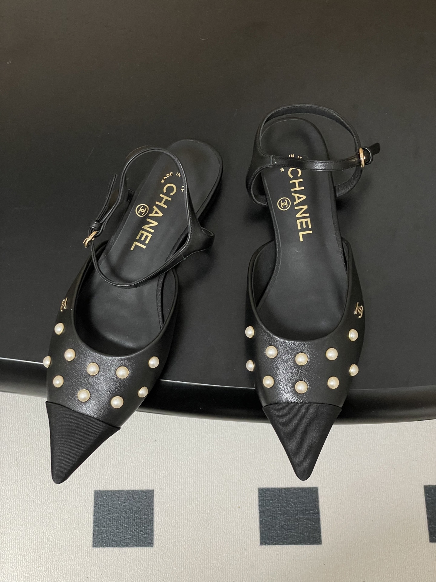 A pair of black Chanel slingback flats with pearl studs, pointed fabric cap toes, and gold CC logo on a black surface. - Hình 2