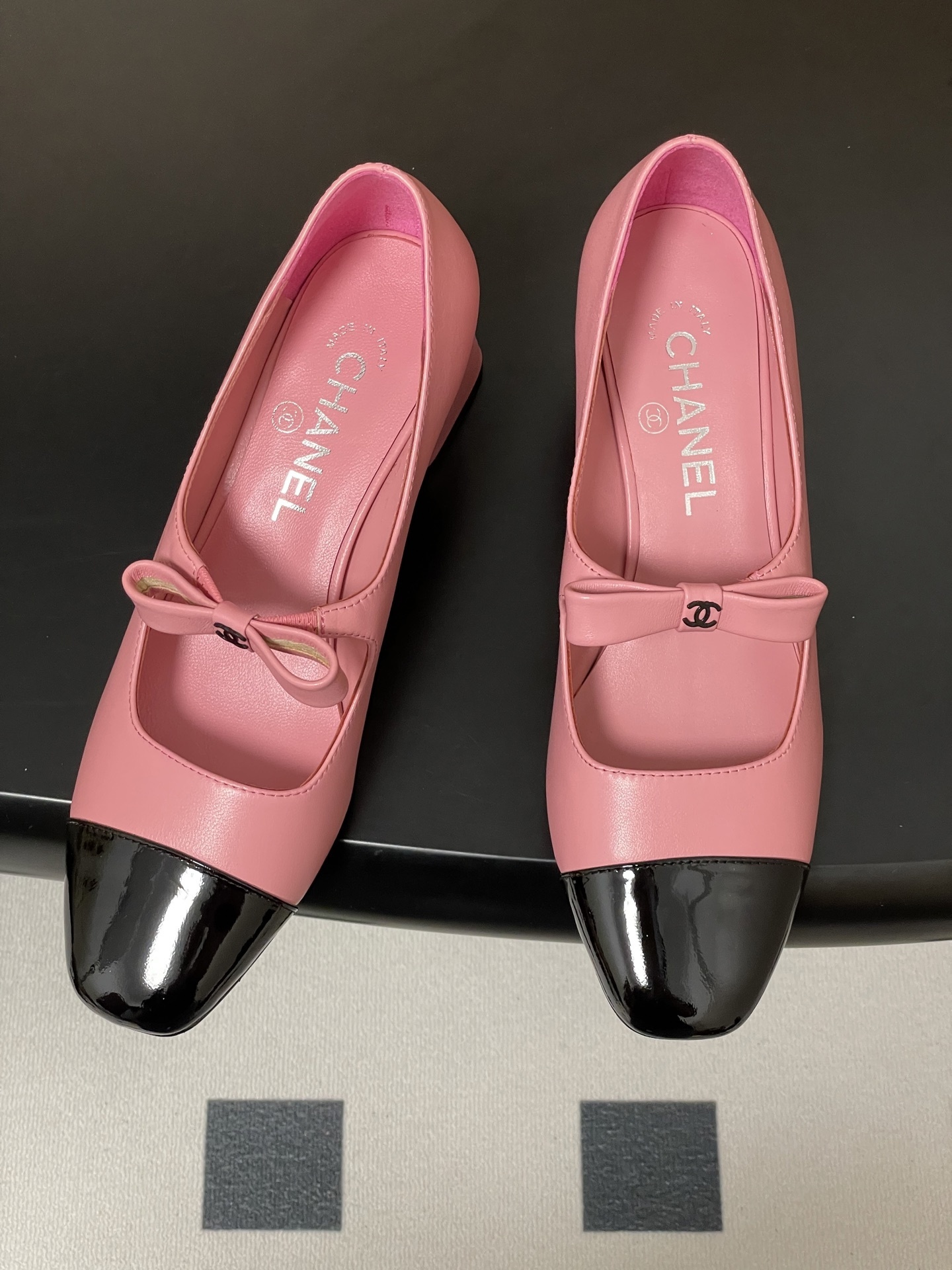 A pair of Chanel pink leather Mary Jane shoes with black patent toe caps and CC logo bows on a black surface - Hình 2
