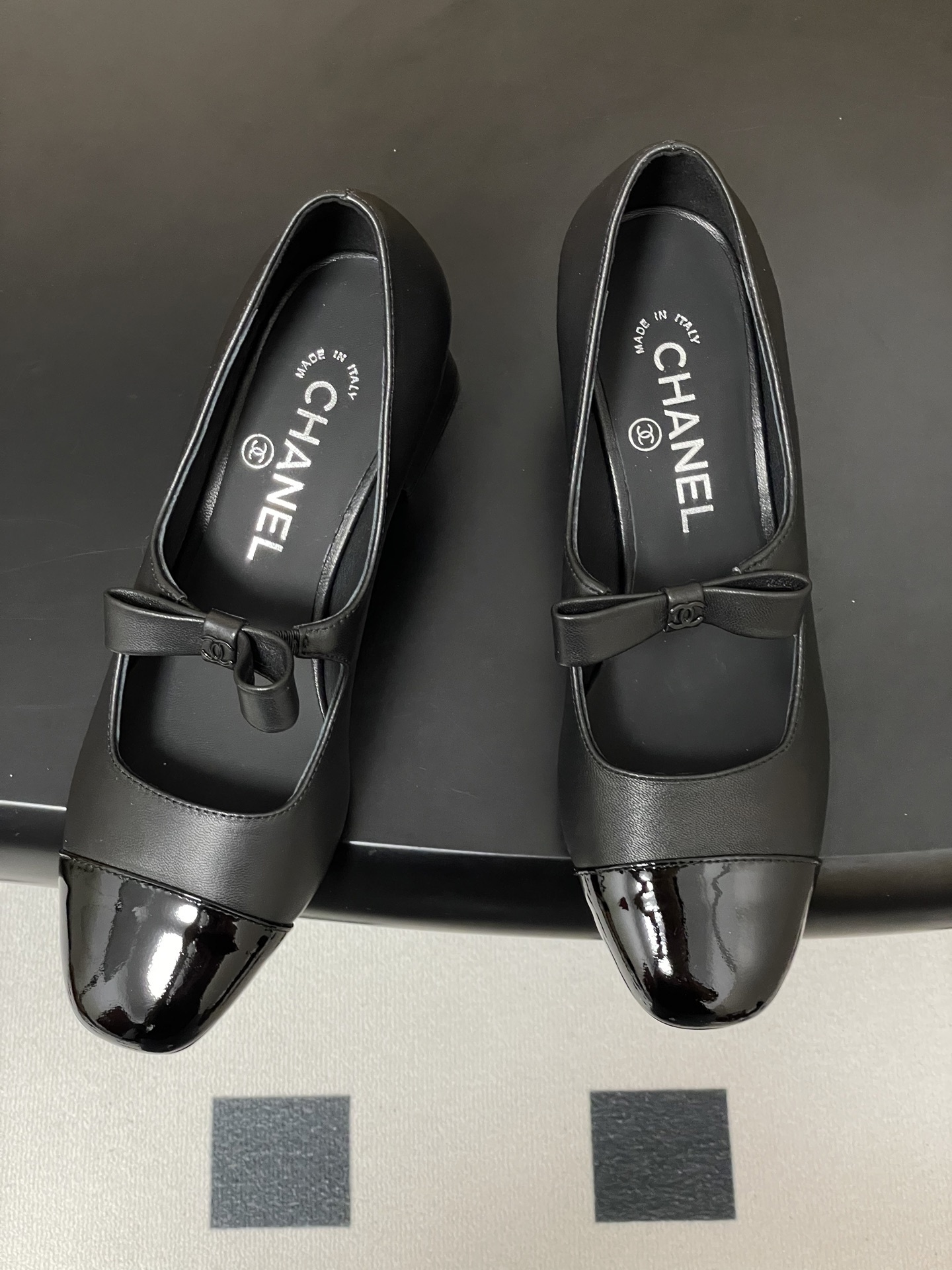 A pair of elegant black Chanel Mary Jane pumps with patent leather cap toes and bow accents featuring the CC logo. - Hình 2