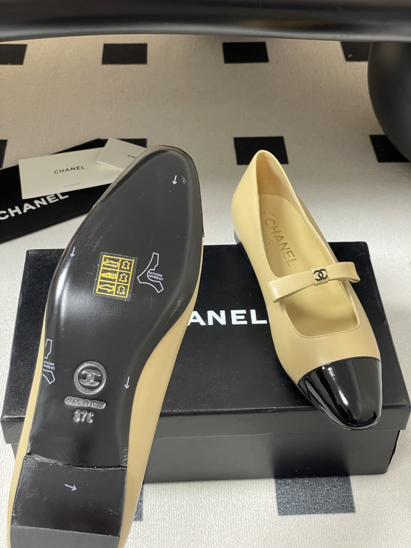 Pair of luxury Chanel beige leather Mary Jane flats with black patent leather cap toes and CC logo straps on a black surface. - Hình 9