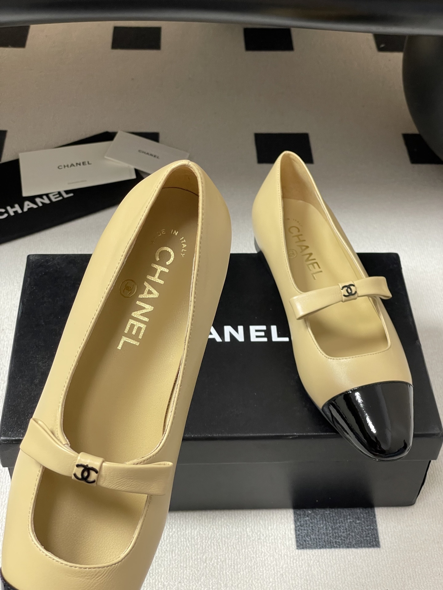 Pair of luxury Chanel beige leather Mary Jane flats with black patent leather cap toes and CC logo straps on a black surface. - Hình 8