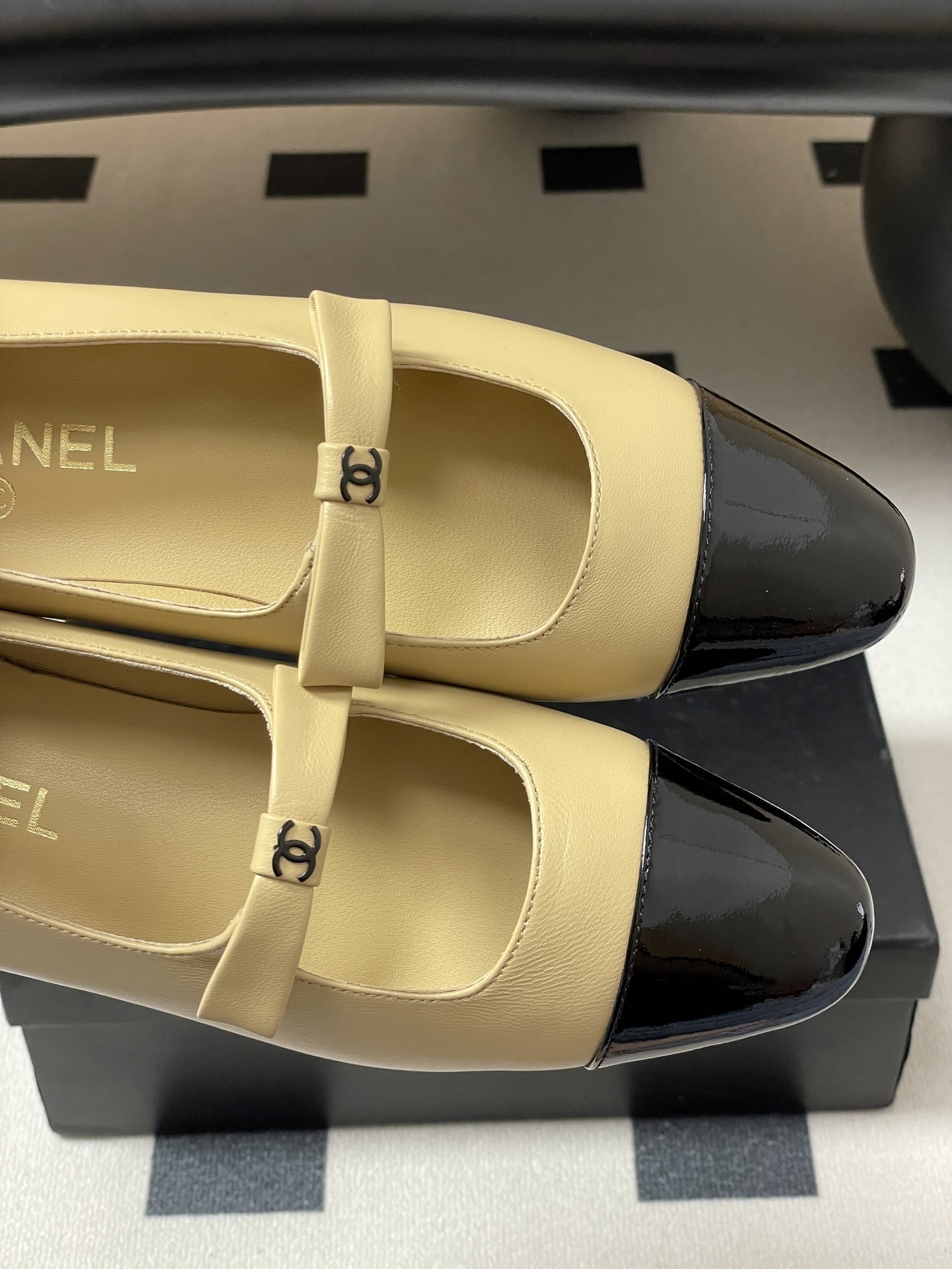Pair of luxury Chanel beige leather Mary Jane flats with black patent leather cap toes and CC logo straps on a black surface. - Hình 7