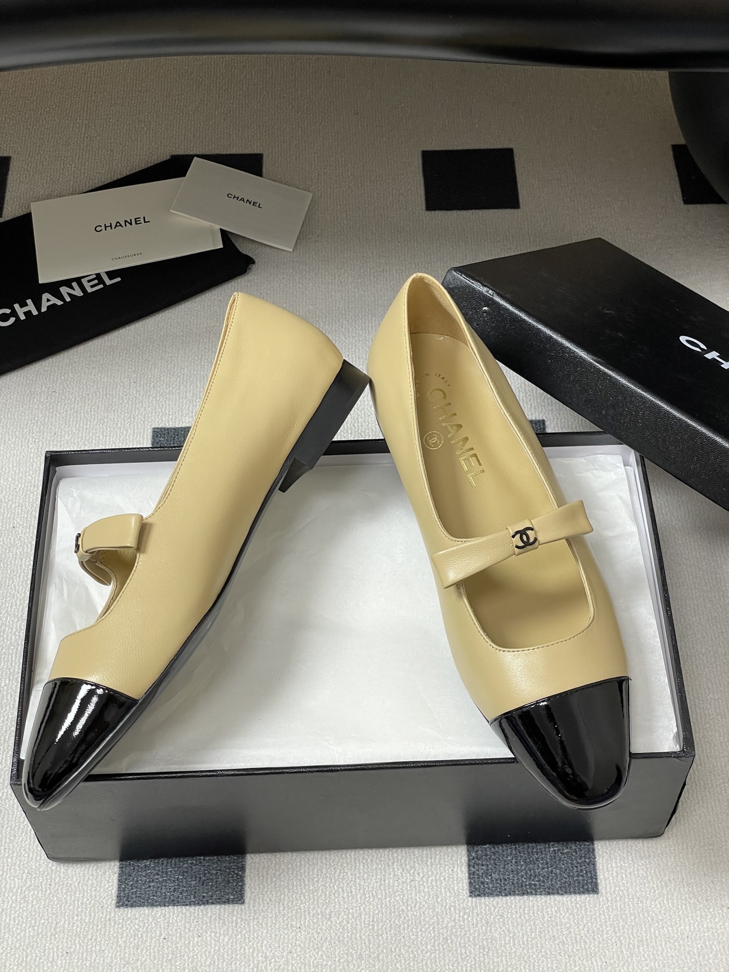 Pair of luxury Chanel beige leather Mary Jane flats with black patent leather cap toes and CC logo straps on a black surface. - Hình 6