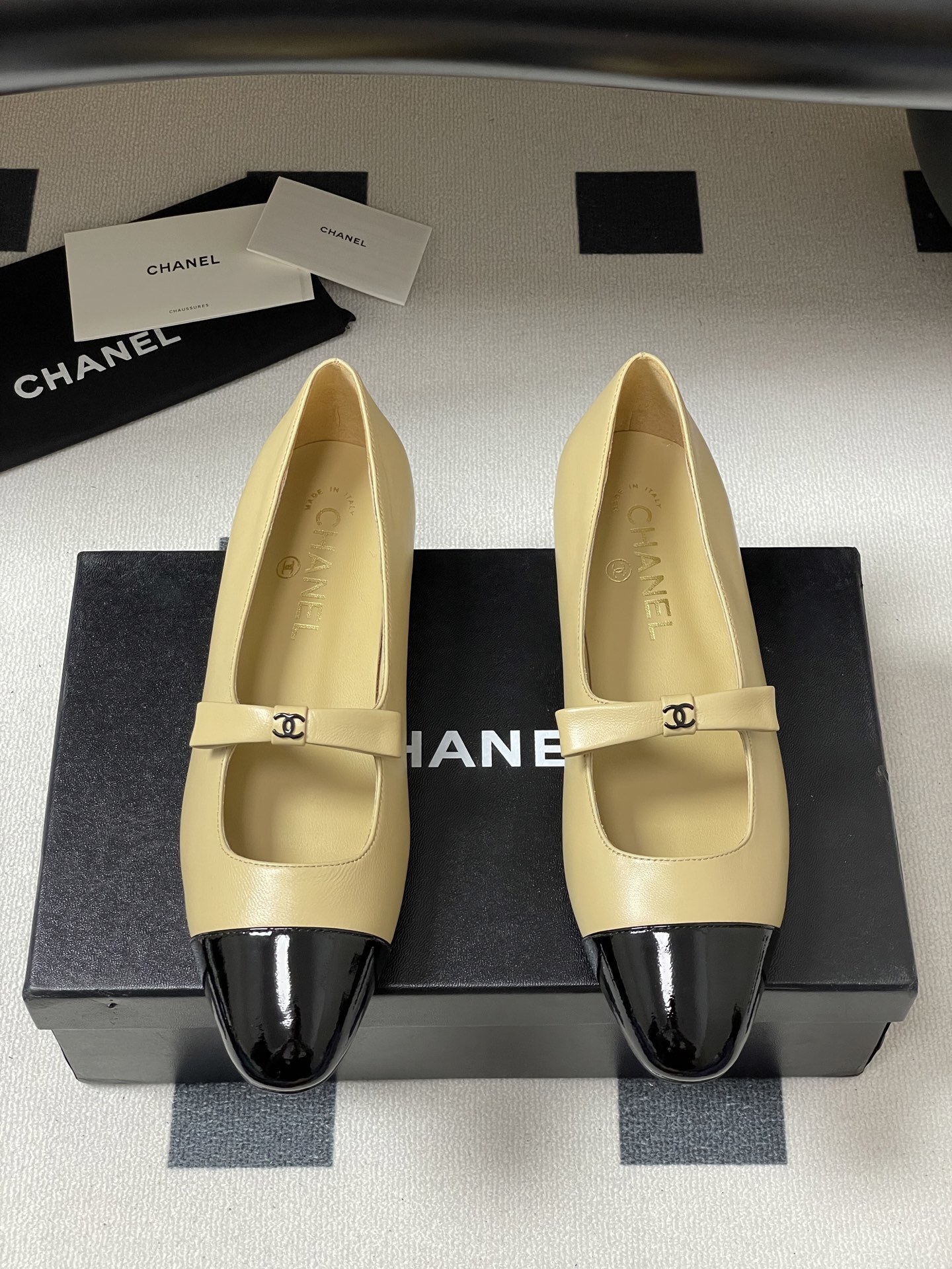 Pair of luxury Chanel beige leather Mary Jane flats with black patent leather cap toes and CC logo straps on a black surface. - Hình 5
