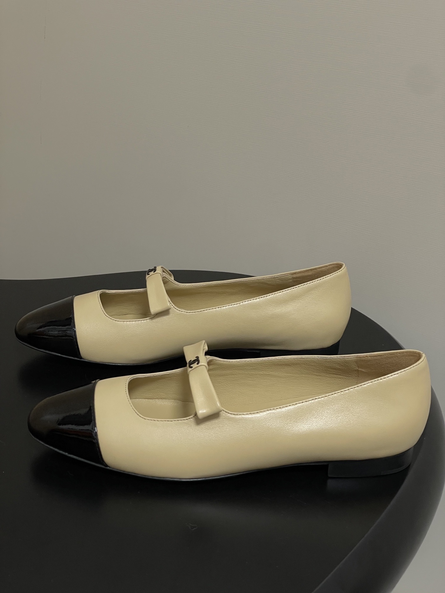 Pair of luxury Chanel beige leather Mary Jane flats with black patent leather cap toes and CC logo straps on a black surface. - Hình 3