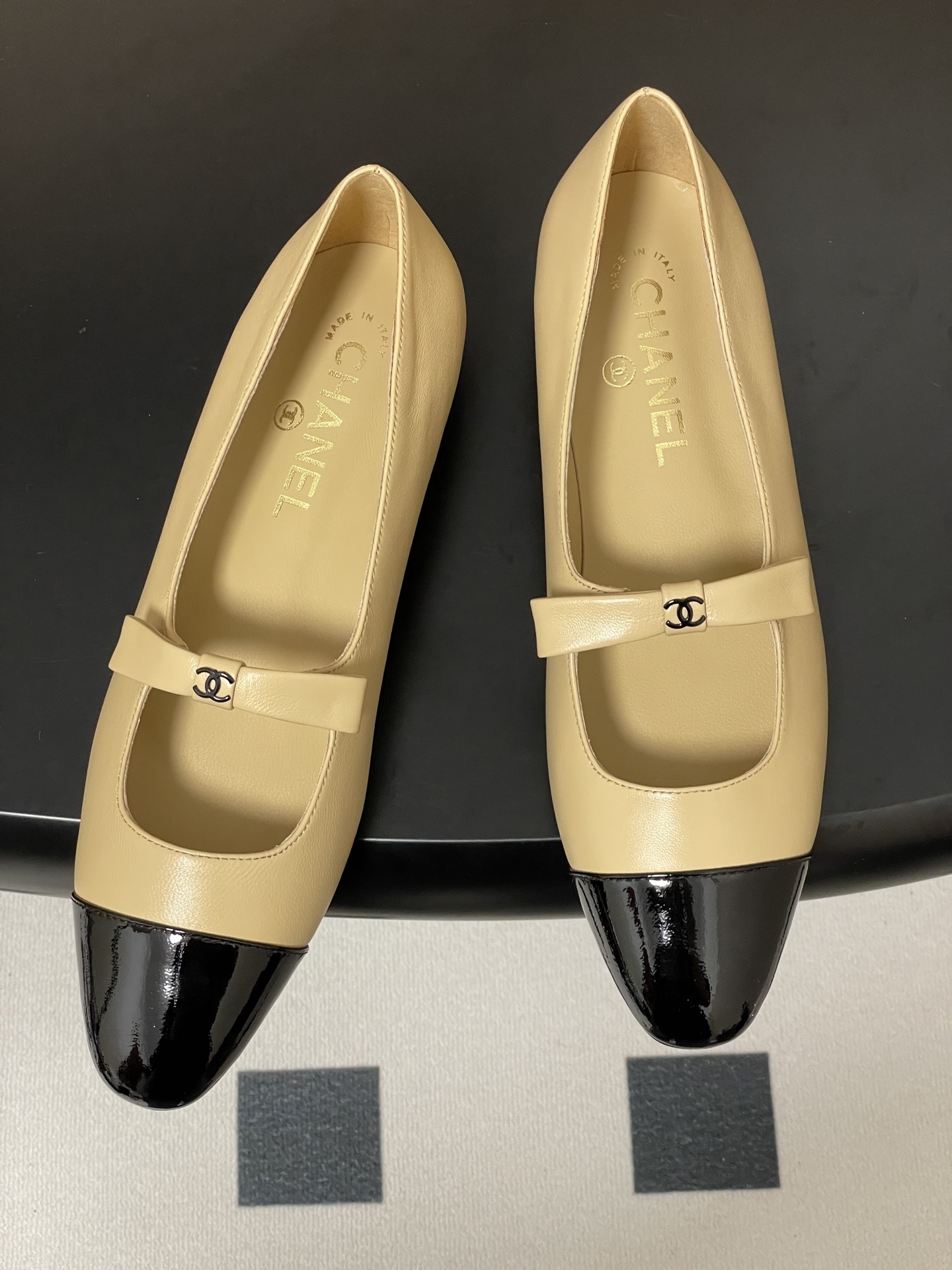 Pair of luxury Chanel beige leather Mary Jane flats with black patent leather cap toes and CC logo straps on a black surface. - Hình 2
