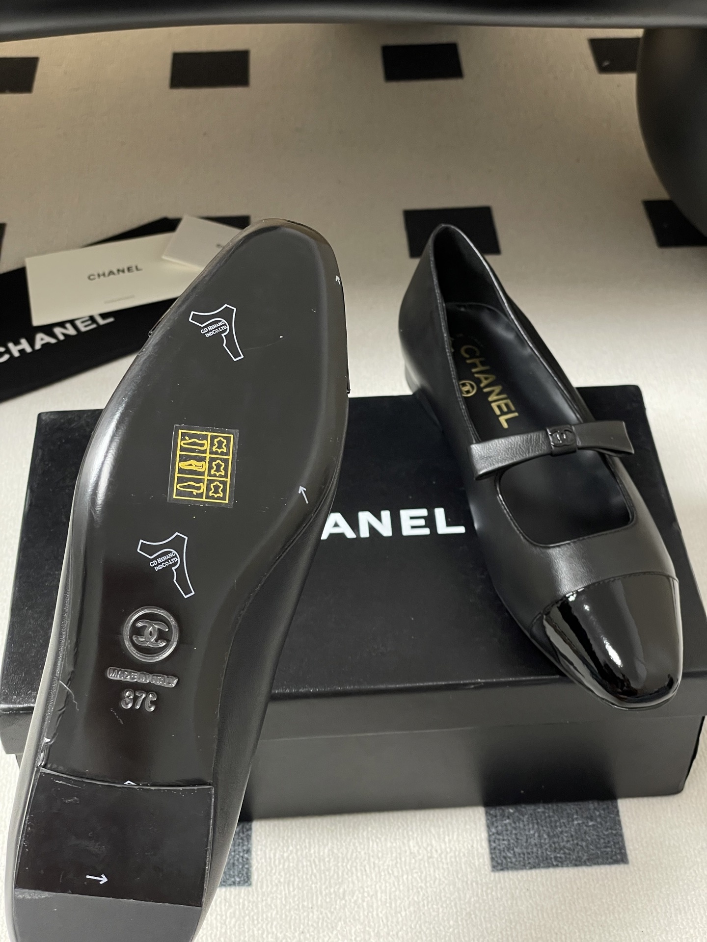 A pair of authentic Chanel black leather Mary Jane flats with glossy patent leather toe caps and CC logo straps displayed on a black surface. - Hình 9